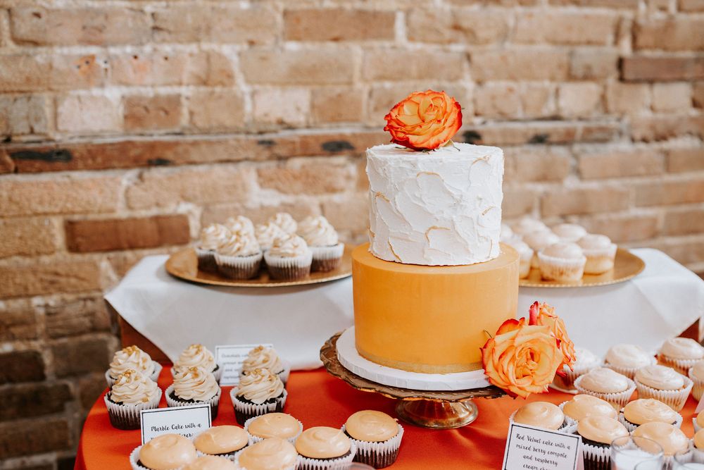 Orange and white wedding dessert table with cakes, cupcakes and treats displayed against exposed brick wall.