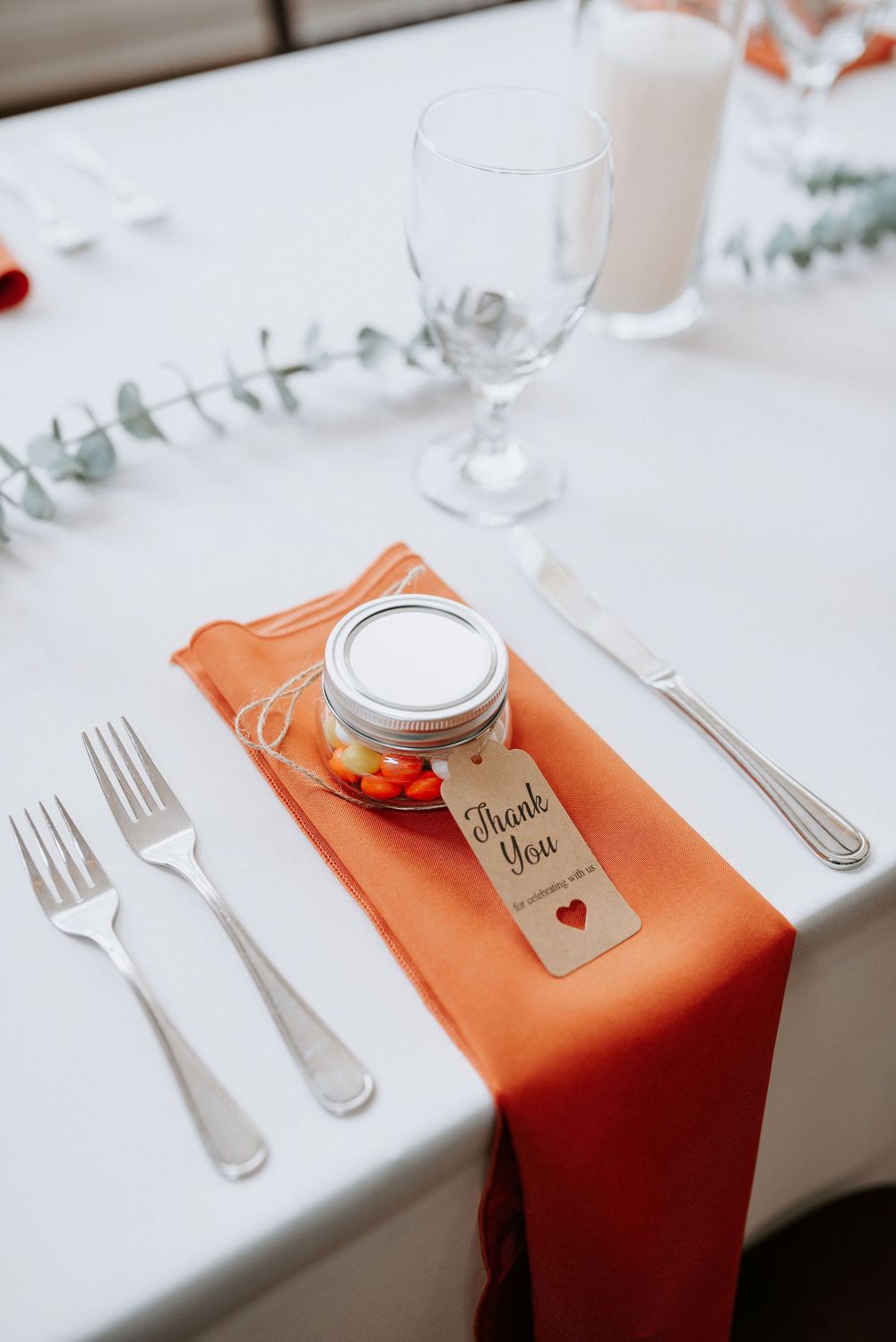 Elegant place settings at a wedding reception with orange napkins and personalized favor jars on white tablecloths.