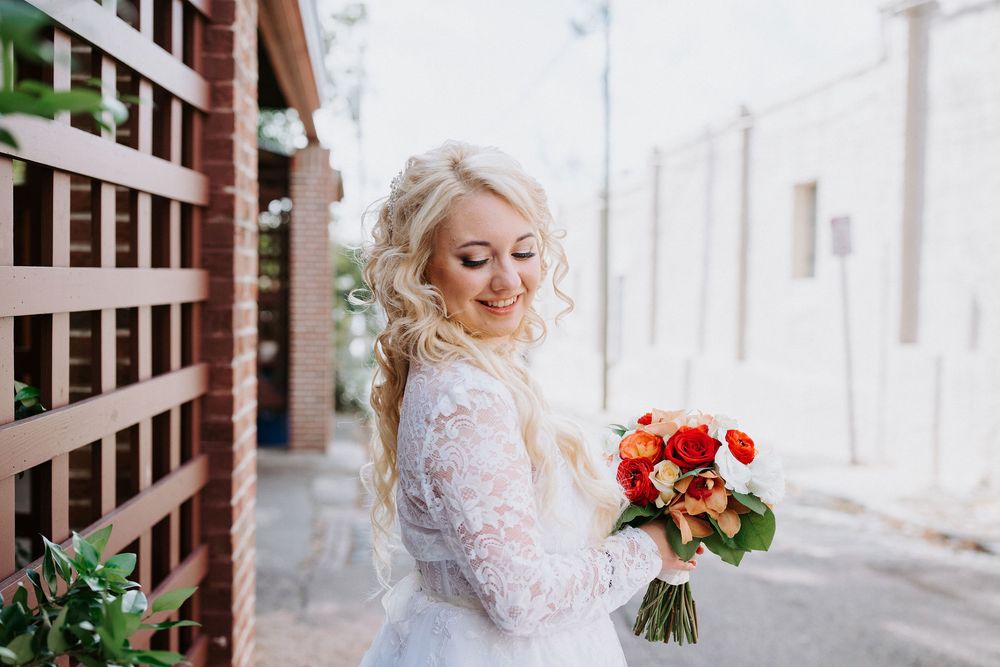 A bride in a white lace gown holds a vibrant red and orange bouquet while standing near wooden railings.