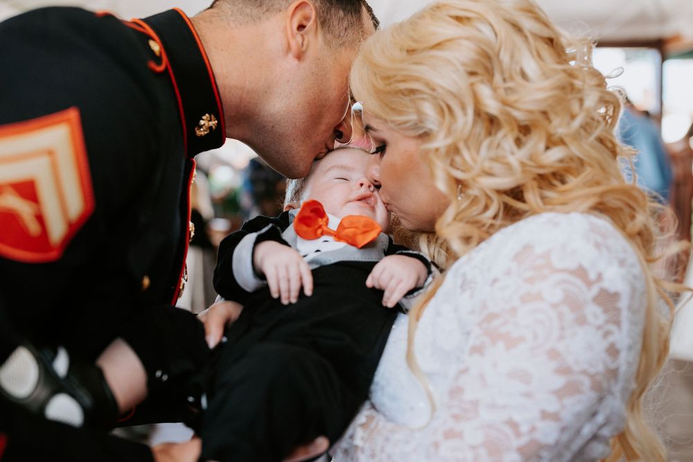 A tender moment between a military service member and partner in formal attire with a baby in dress clothes between them.