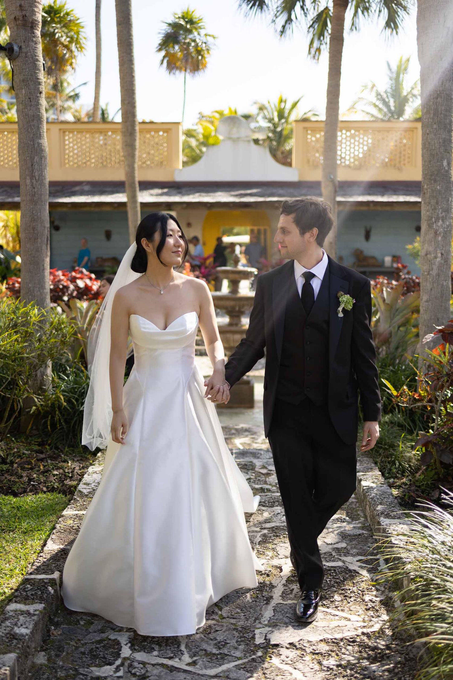 A bride and groom walk together through a tropical garden path lined with palm trees on their wedding day.