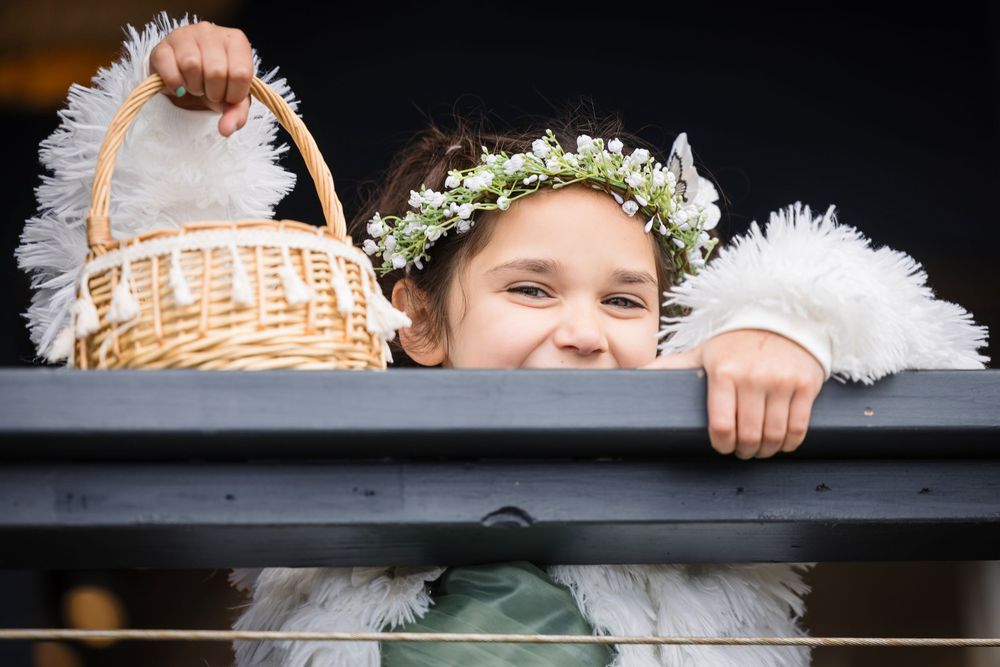 A flower girl in a white dress and floral crown peeks over a wooden railing at a wedding ceremony.
