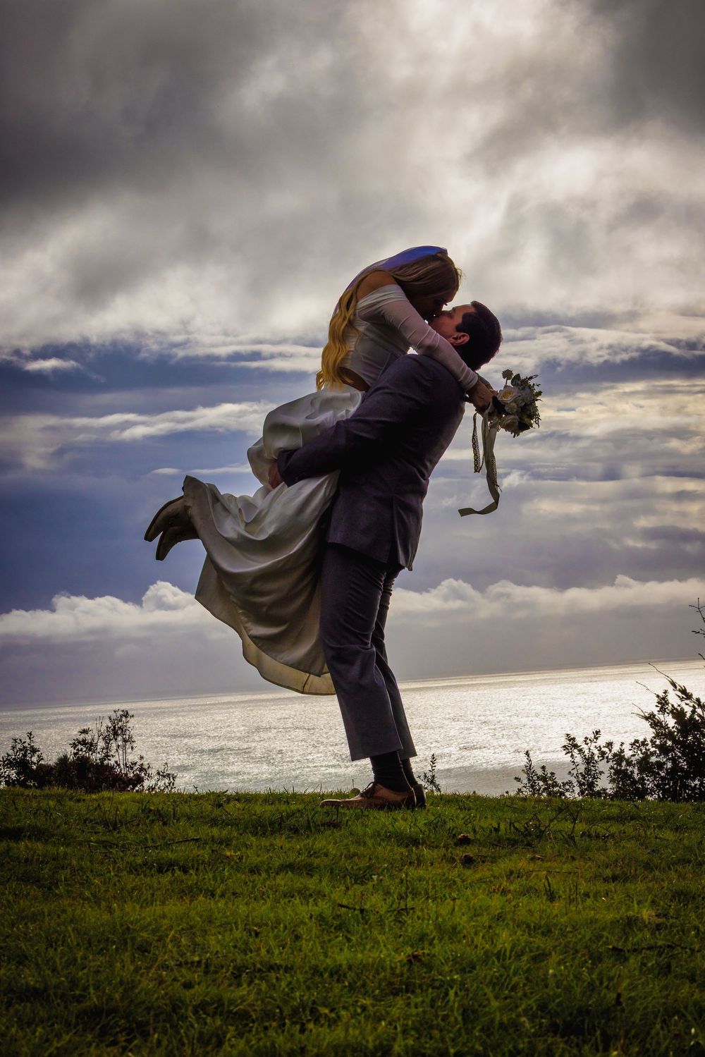 Romantic couple embracing on hilltop against dramatic cloudy sky and ocean view at sunset.