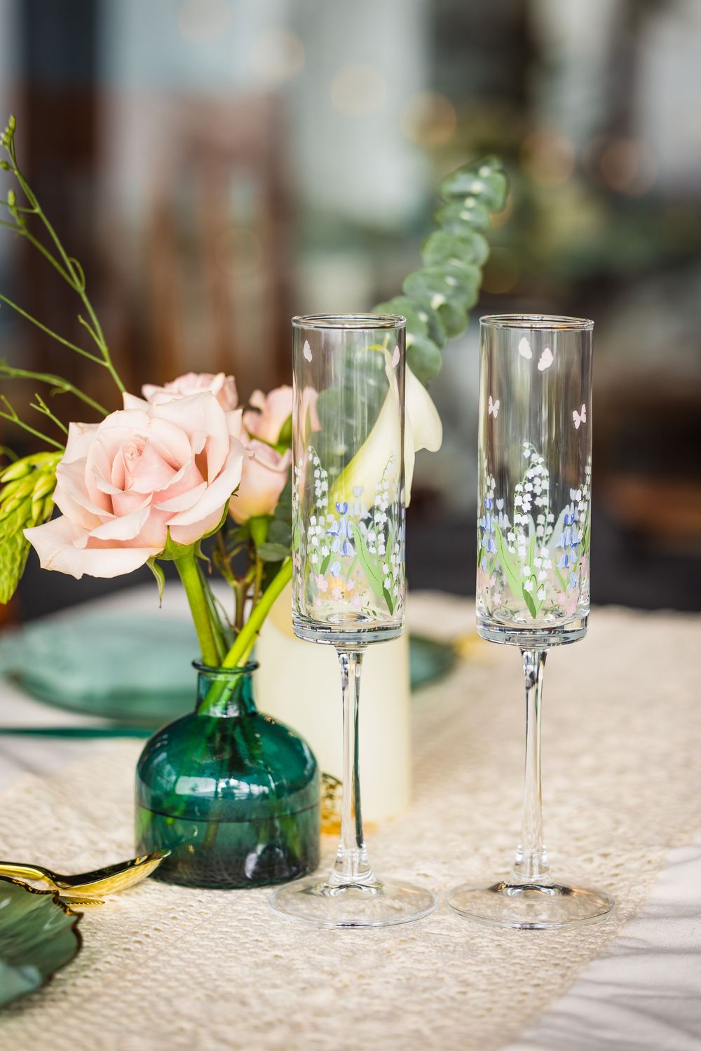 Elegant table setting with green glass vases holding pink roses and crystal wine glasses on textured linen tablecloth.