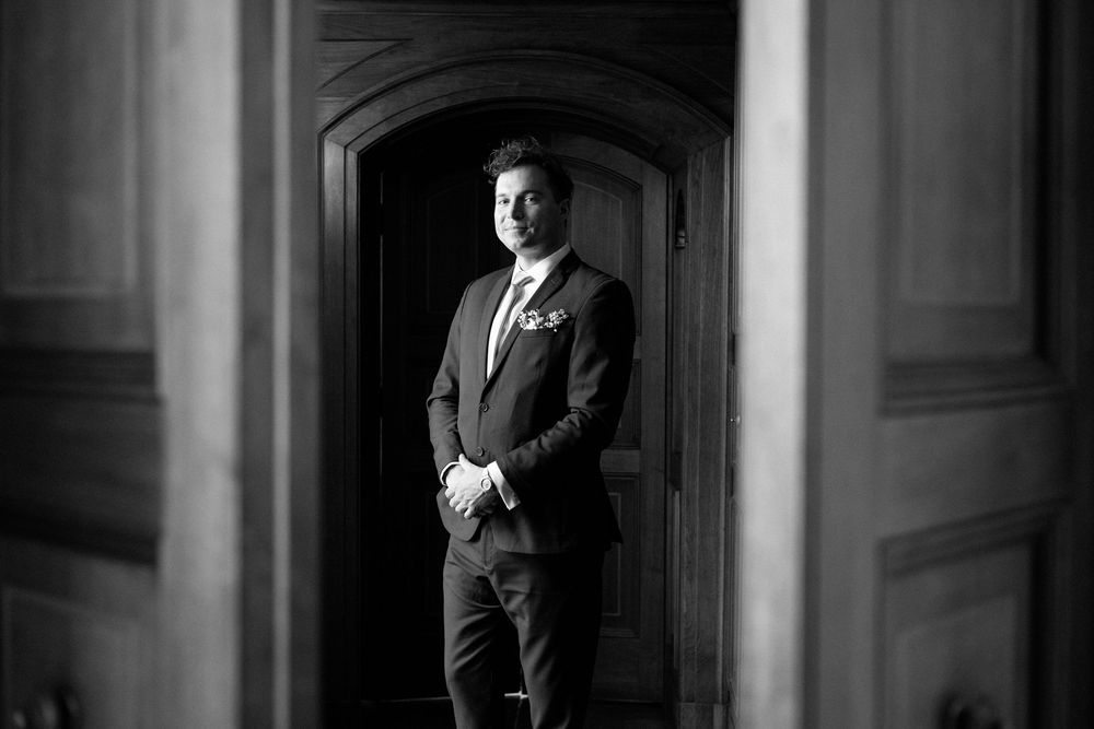 Dramatic black and white photo of a groom standing in a doorway with classic architectural details.