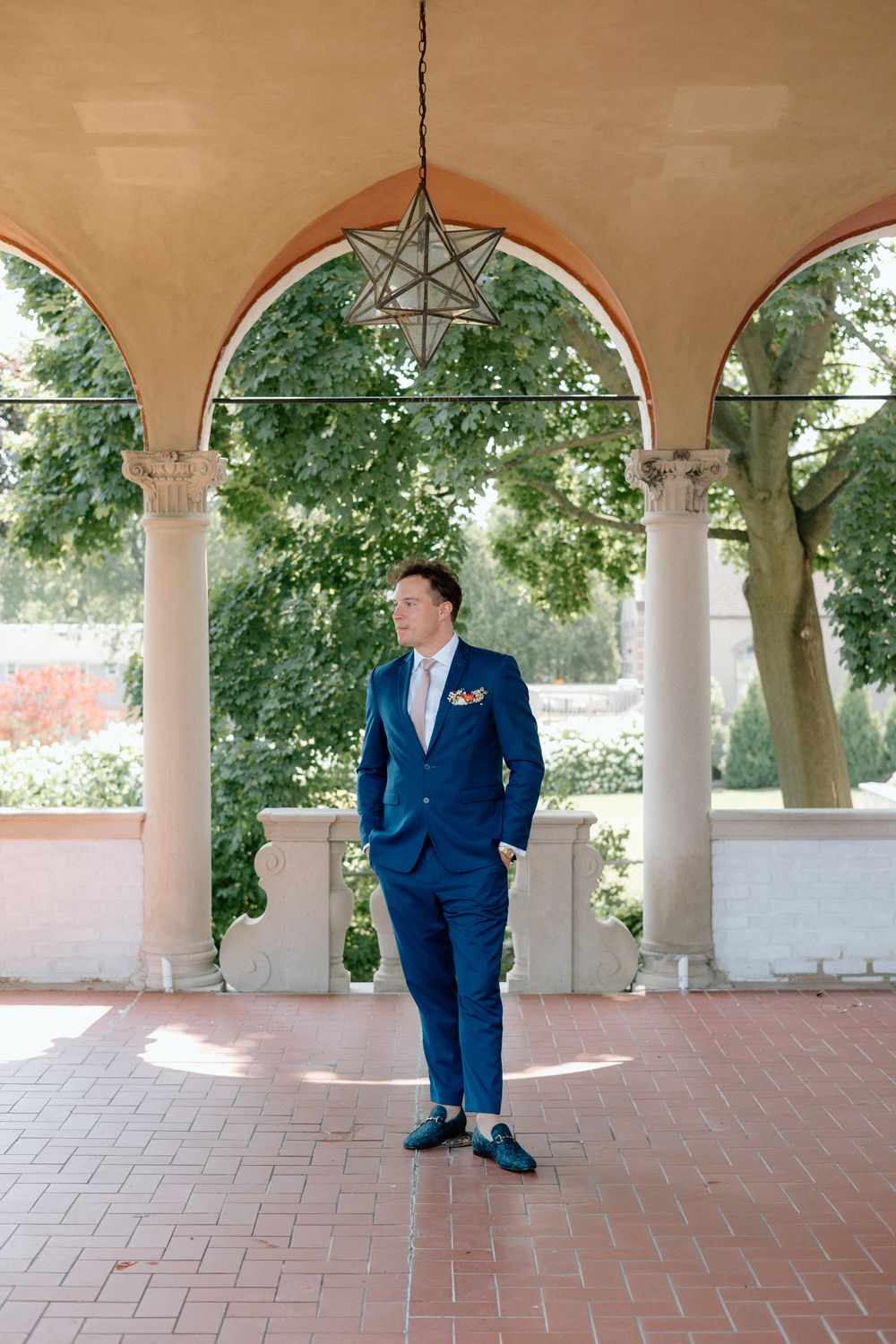 Groom in navy blue suit poses under elegant archways with stone columns on a terracotta tiled patio.