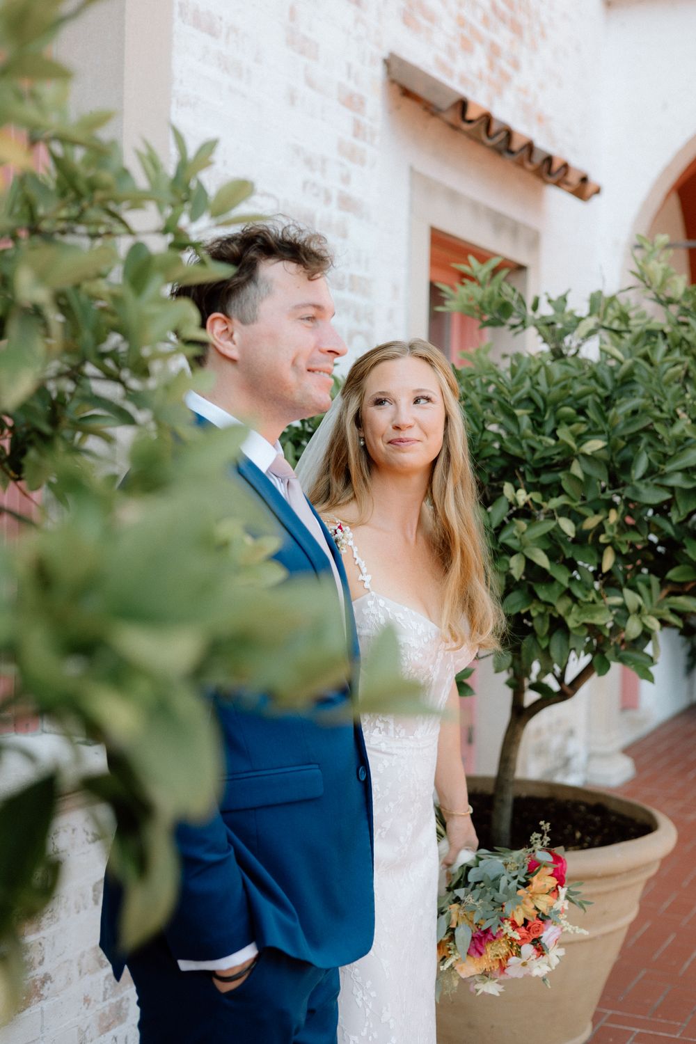 A bride in a white dress and groom in a navy suit share romantic moments in a garden setting with lush greenery.