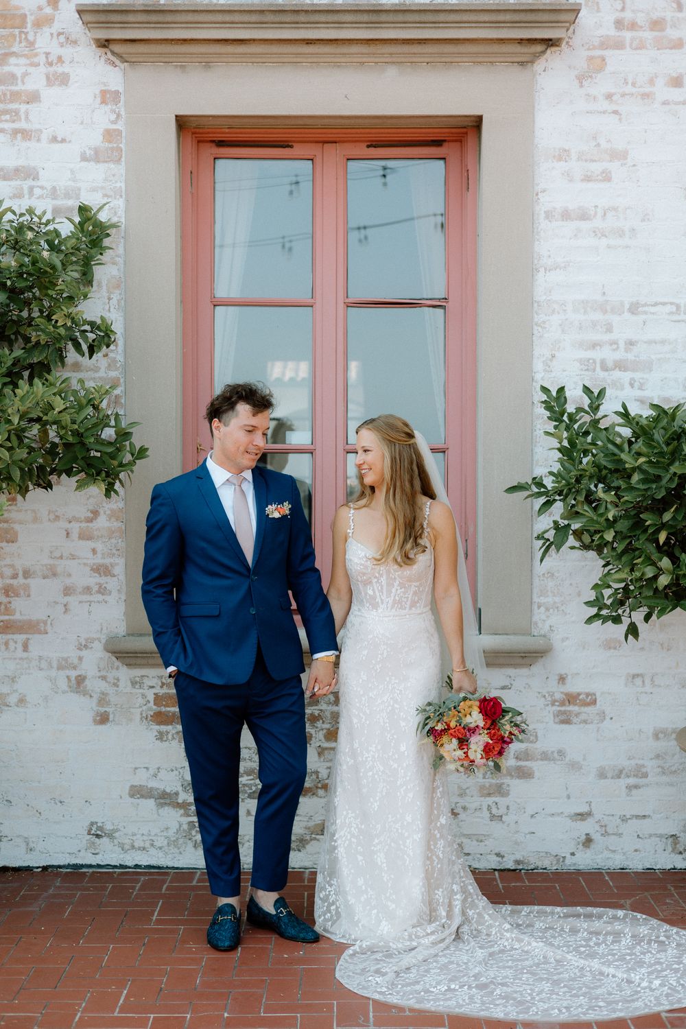 Bride and groom in formal attire pose against a white brick wall with coral doors.