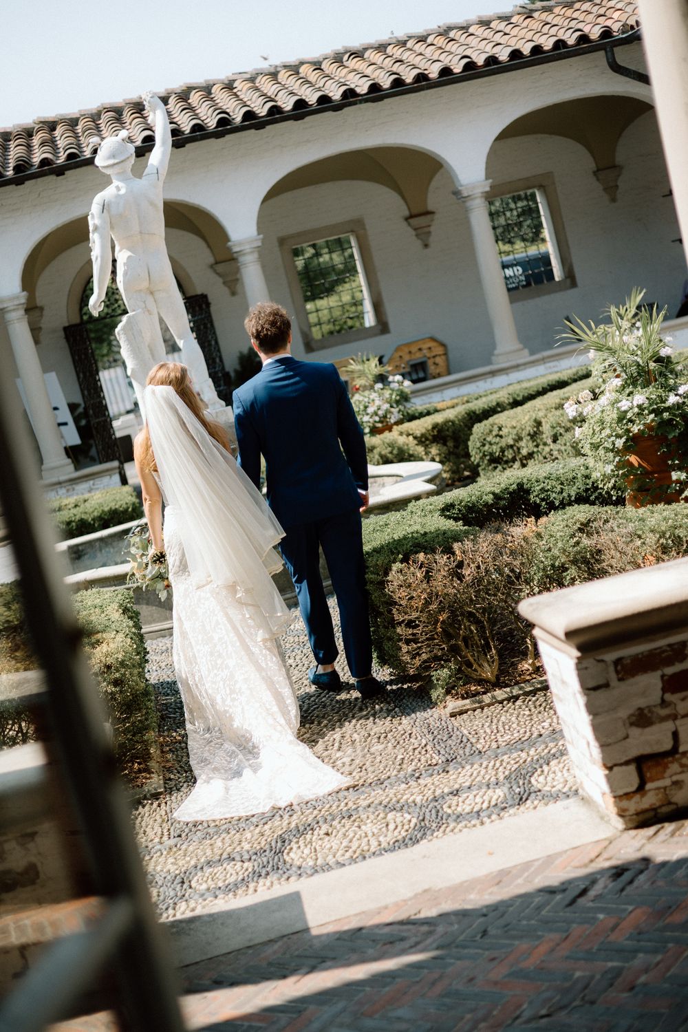 A wedding couple walks along a Spanish colonial style courtyard with arched columns and terra cotta tiles.