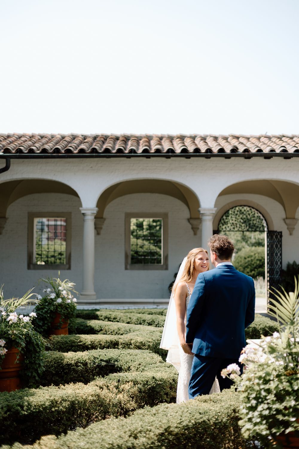 Romantic couple embraces in an elegant Spanish-style courtyard with white arched columns and lush gardens.