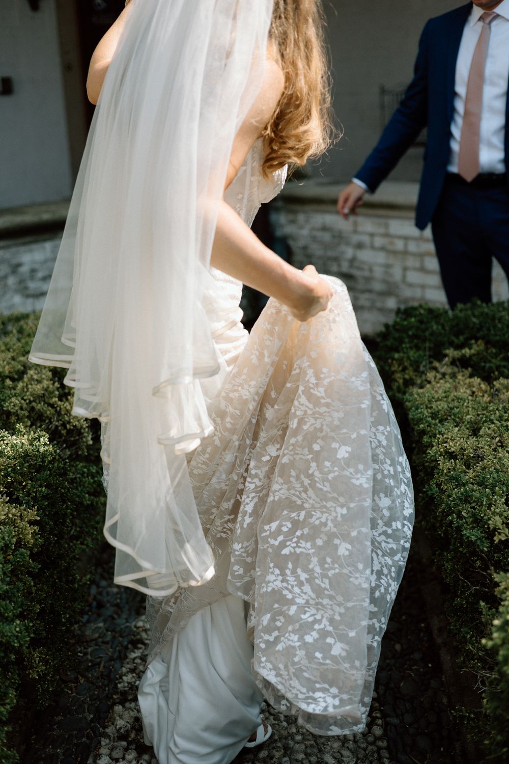Close-up detail of a bride's delicate lace wedding dress and flowing veil against greenery.