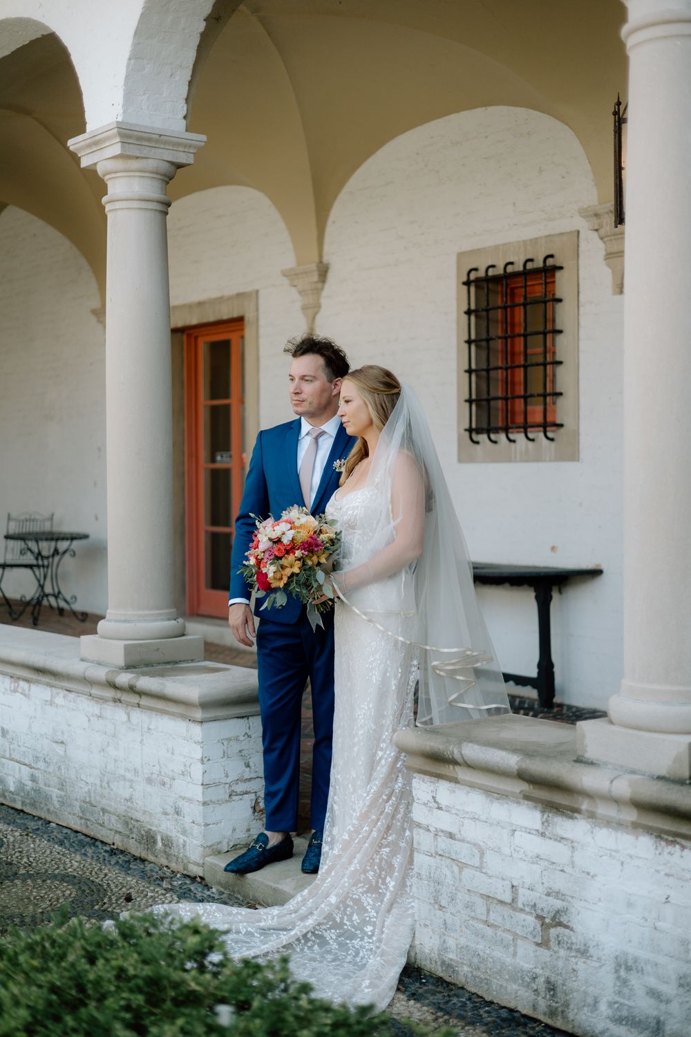 A romantic wedding couple shares intimate moments under a white columned archway at a historic mission-style venue.
