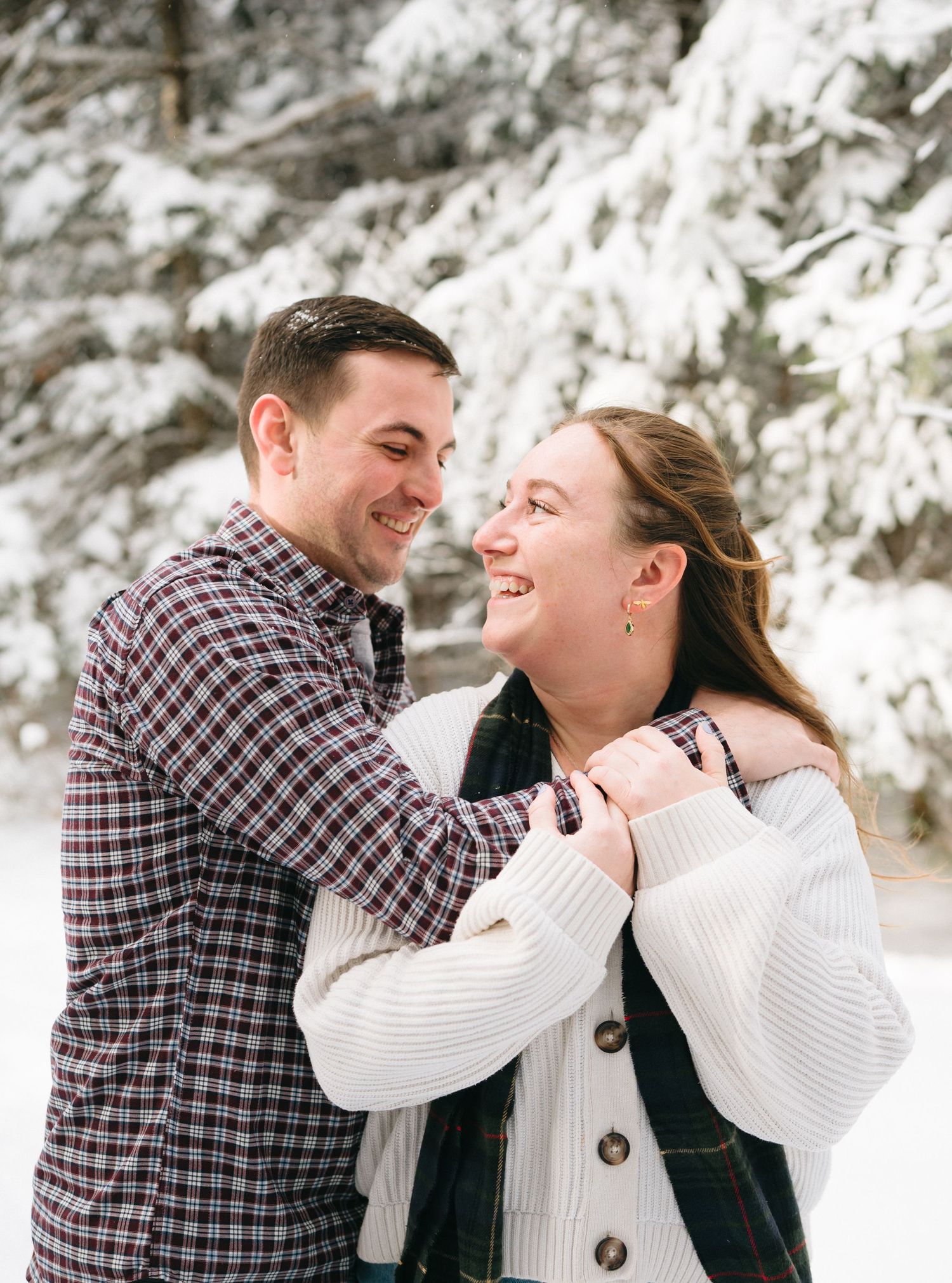 A couple shares joyful laughter while embracing in a snowy winter setting wearing cozy sweaters.