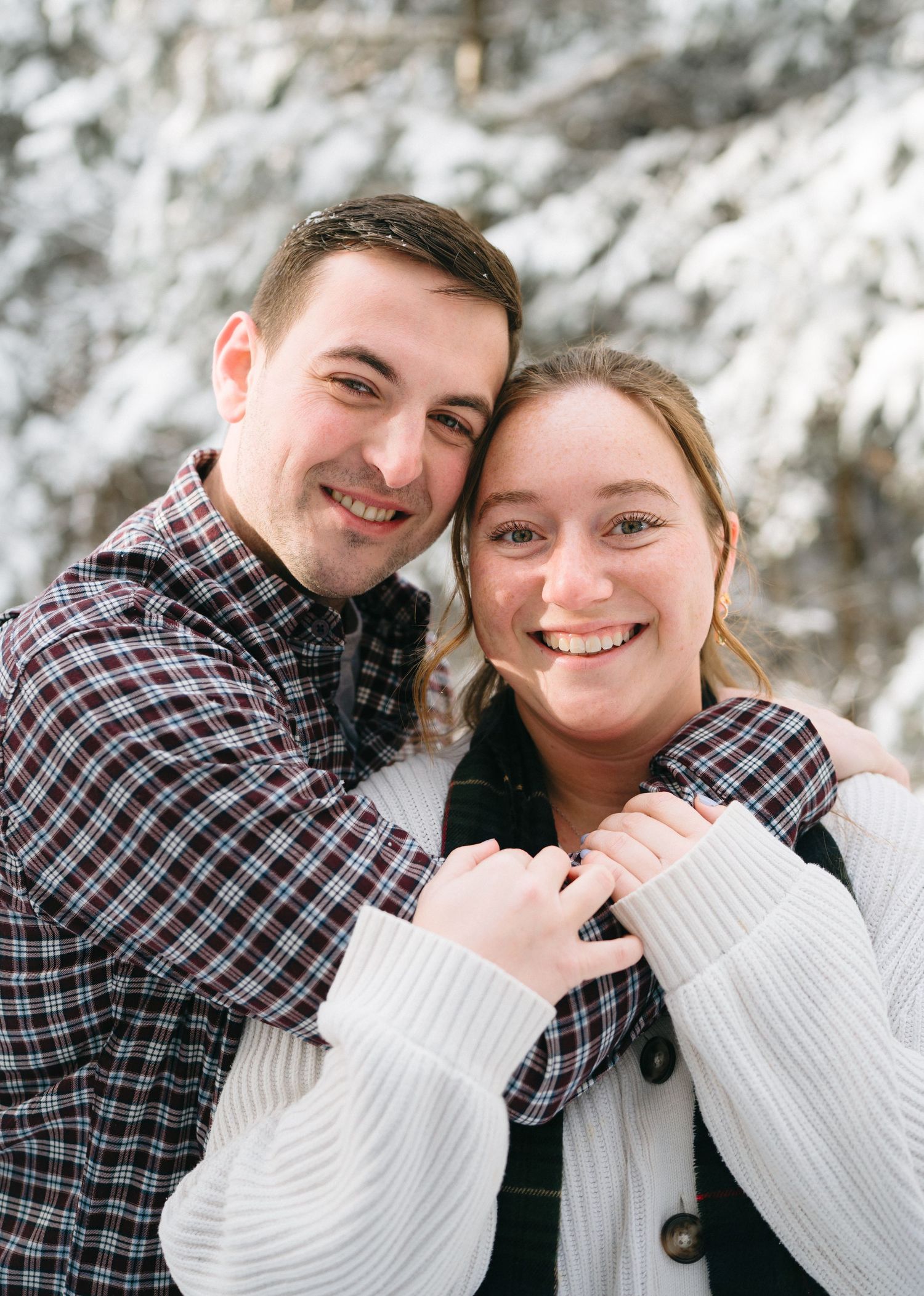 A couple wearing matching plaid shirts and white sweaters pose together outdoors with snowy scenery in the background.