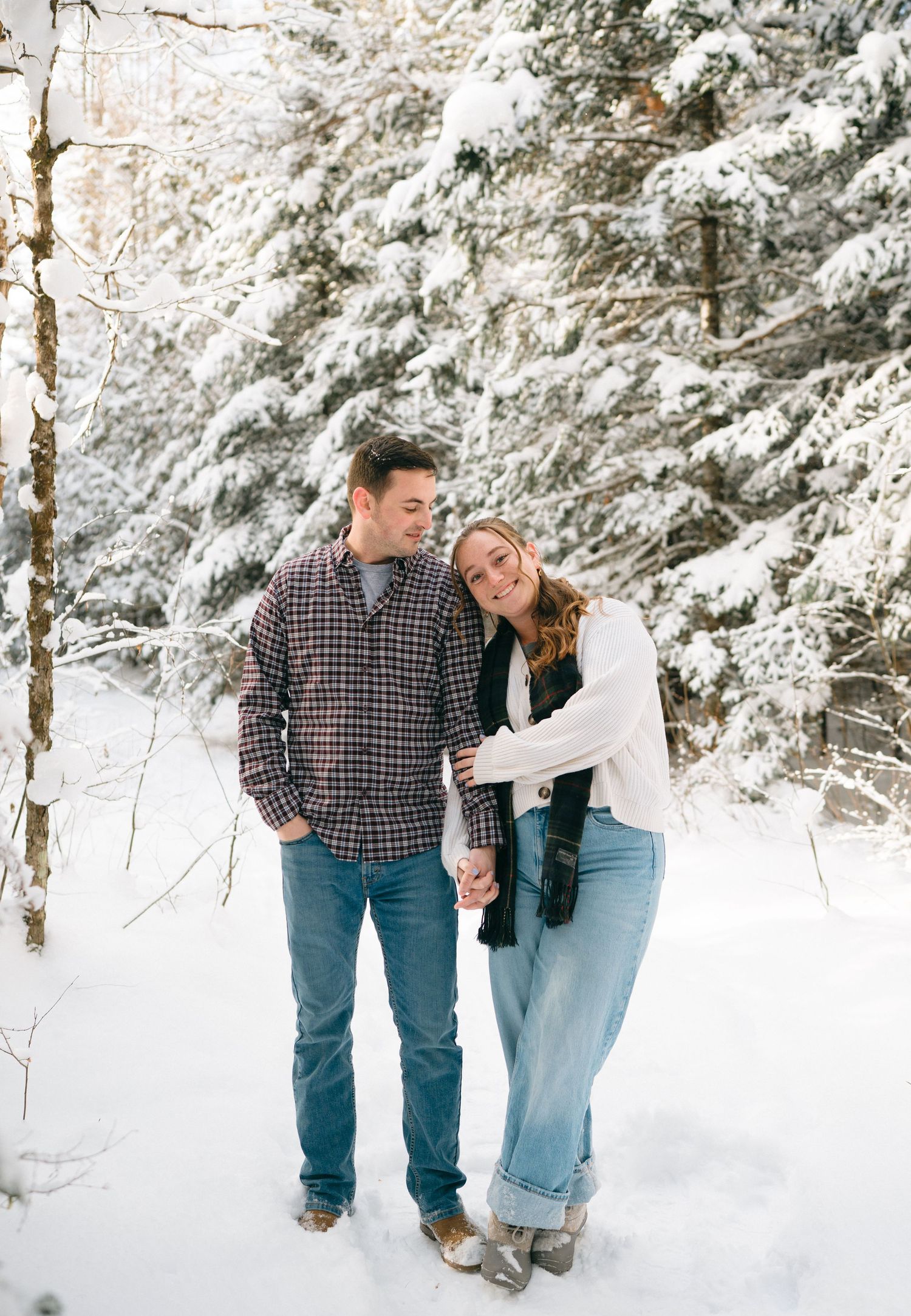 A couple stands close together in a snowy winter forest with snow-covered evergreen trees in the background.