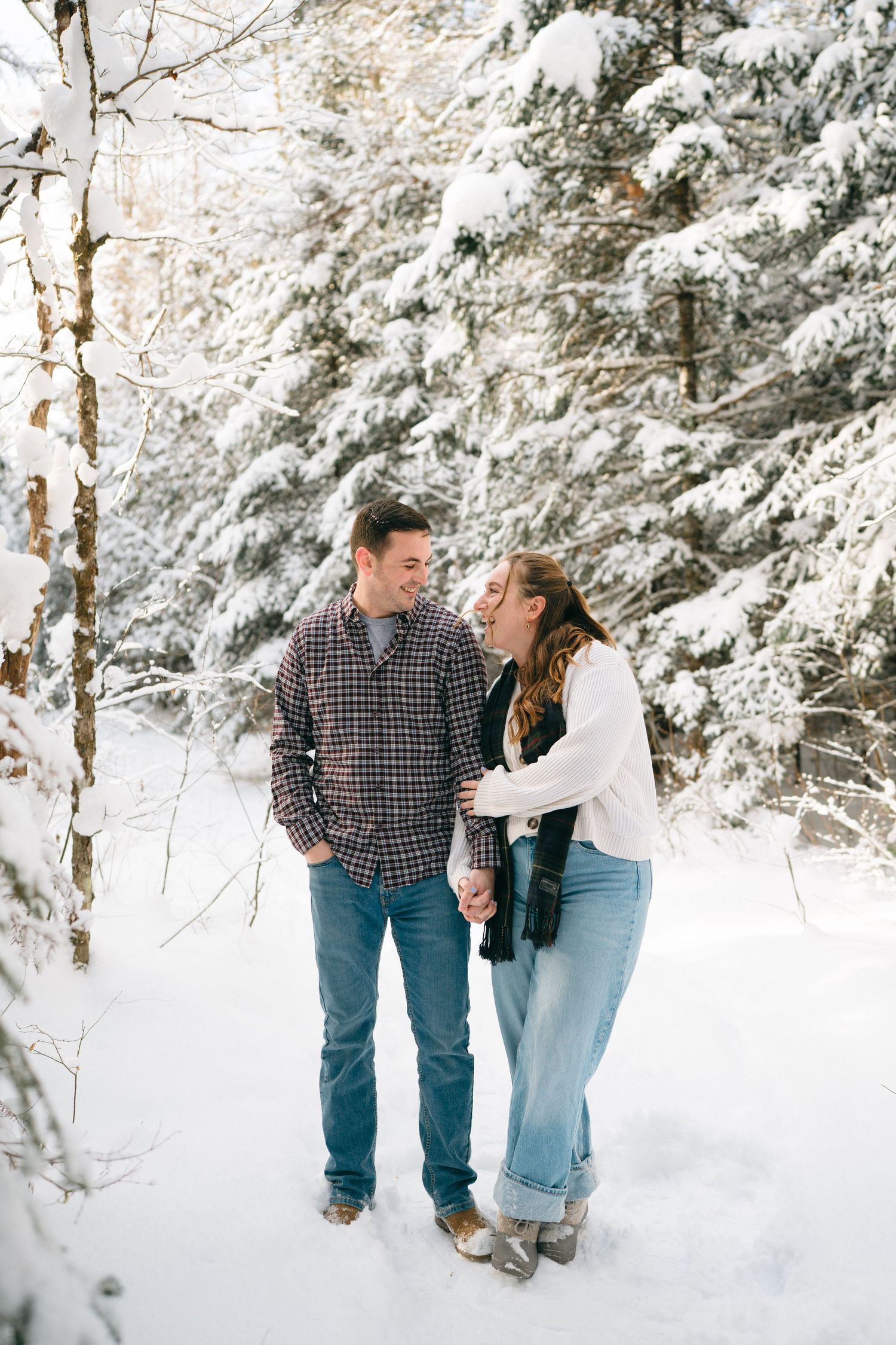 A couple shares a joyful moment together in a snowy winter forest setting with snow-covered evergreen trees.