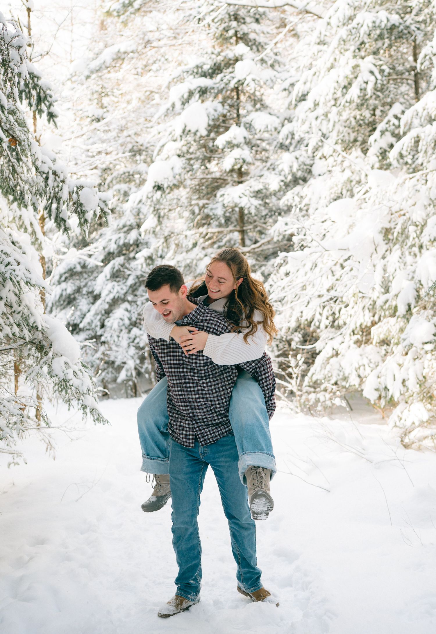 A playful piggyback ride moment between a couple in a snowy forest setting surrounded by frost-covered trees.