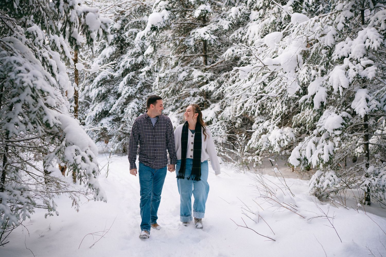 A couple walks hand in hand through a snowy forest path surrounded by snow-covered evergreen trees.
