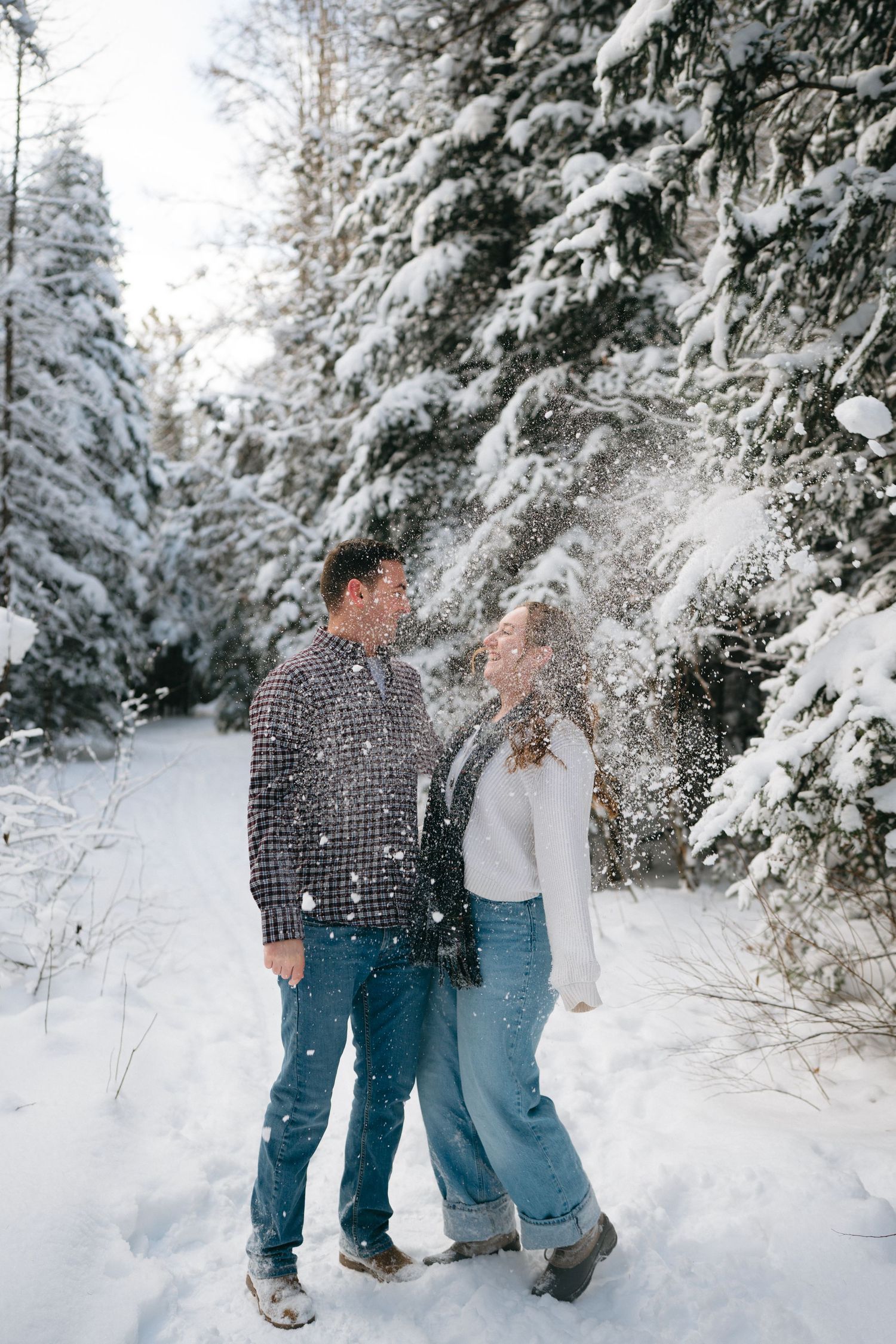 A couple plays together in a snow-covered forest path surrounded by evergreen trees dusted with fresh white snow.