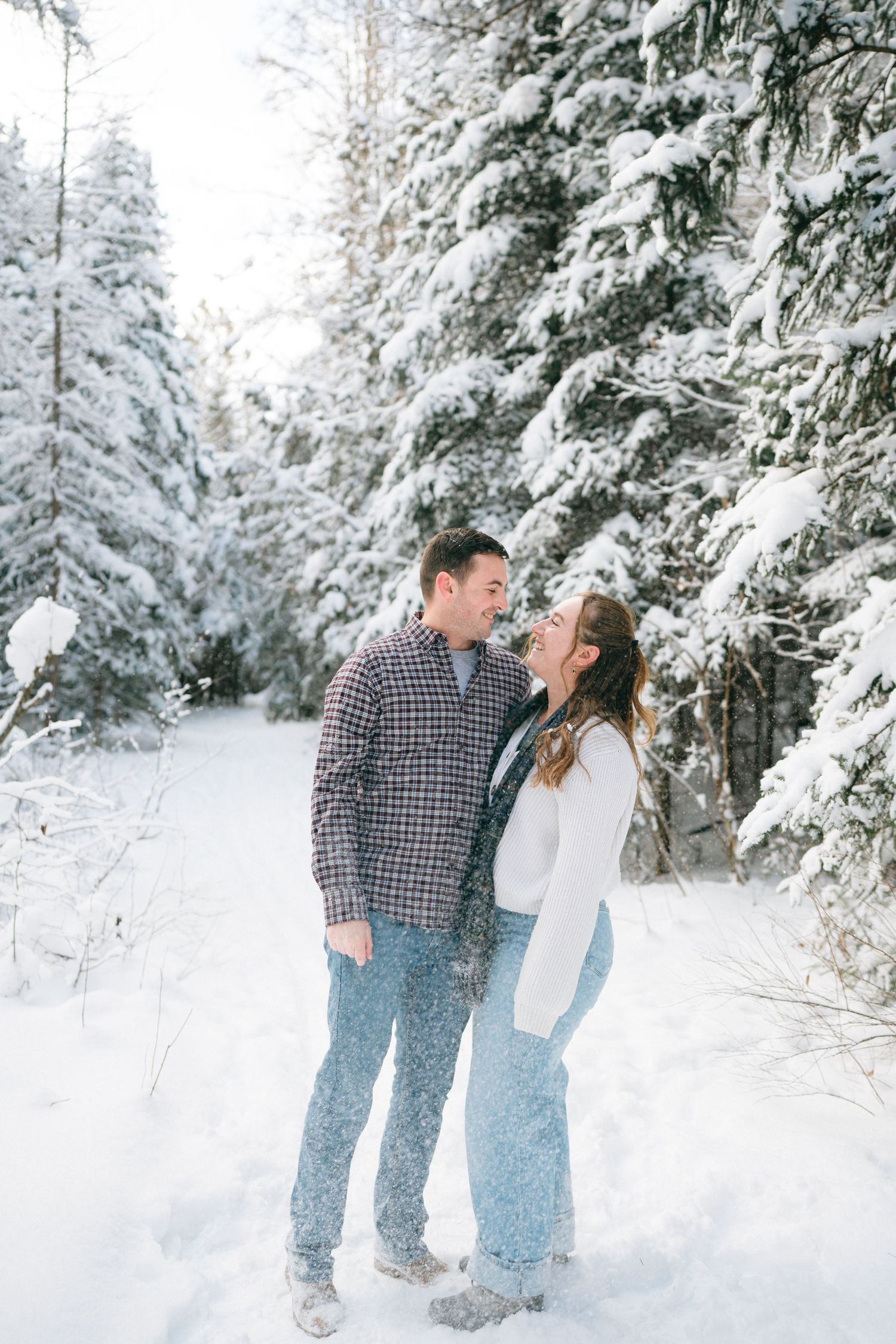 A couple shares a joyful moment together in a snowy winter forest landscape with snow-covered evergreen trees.