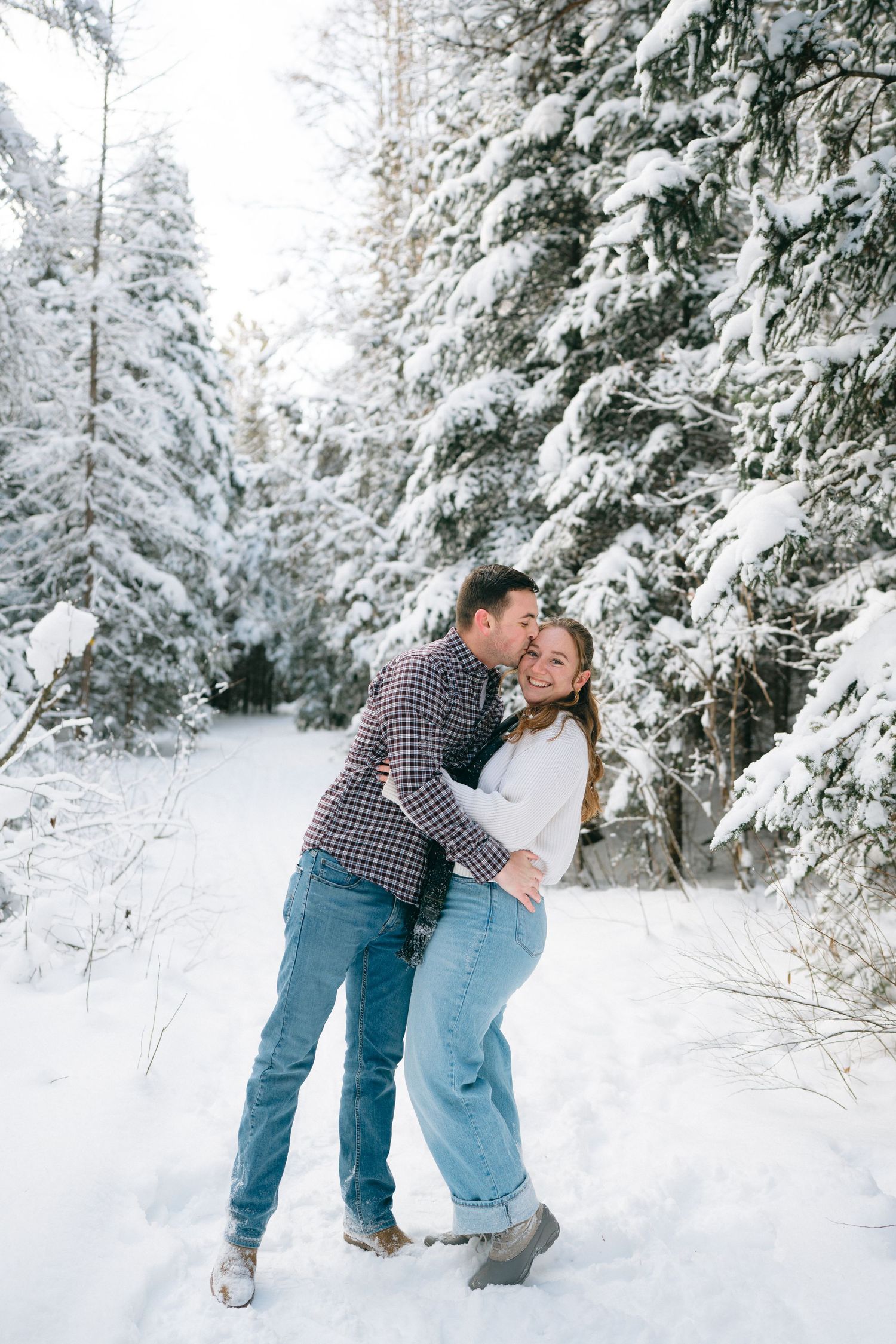 A couple embraces in a snowy winter forest setting with snow-covered evergreen trees in the background.