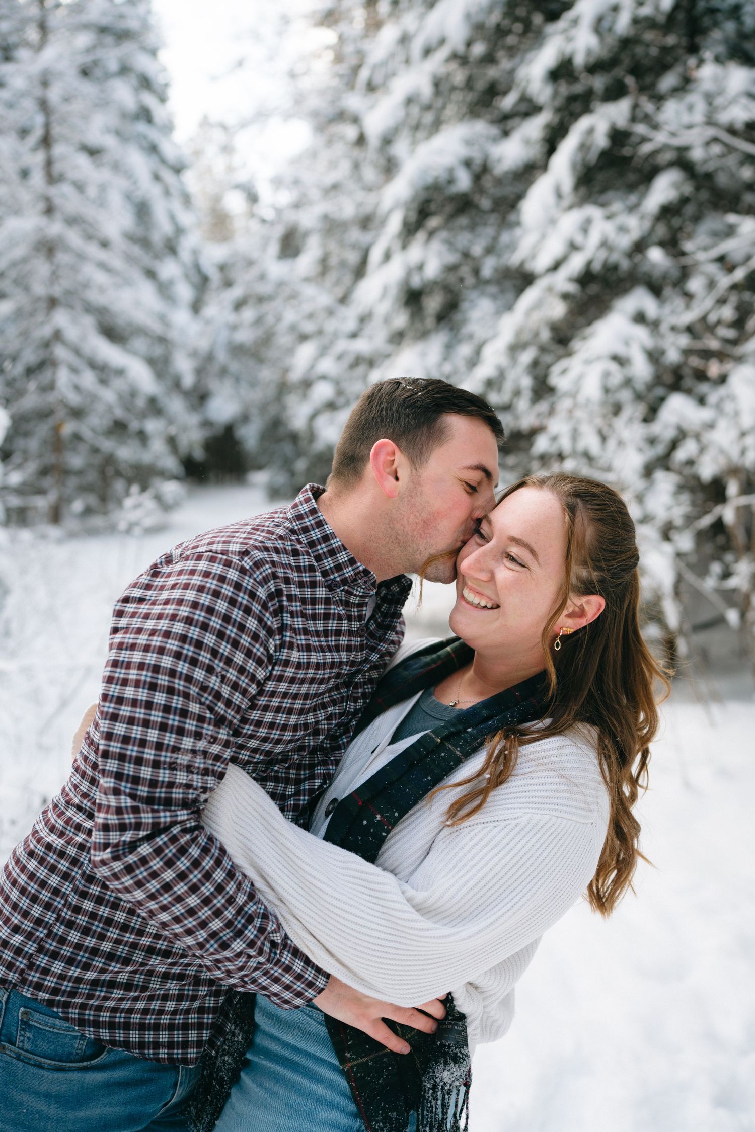 A couple shares a romantic moment in a snowy winter forest setting, surrounded by snow-covered evergreen trees.