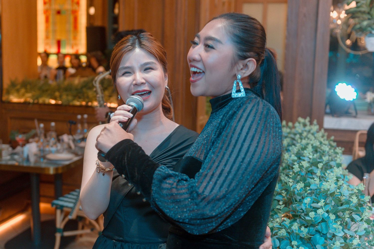 Two people in dark outfits sing together with microphones at an indoor event with decorative lighting and greenery.