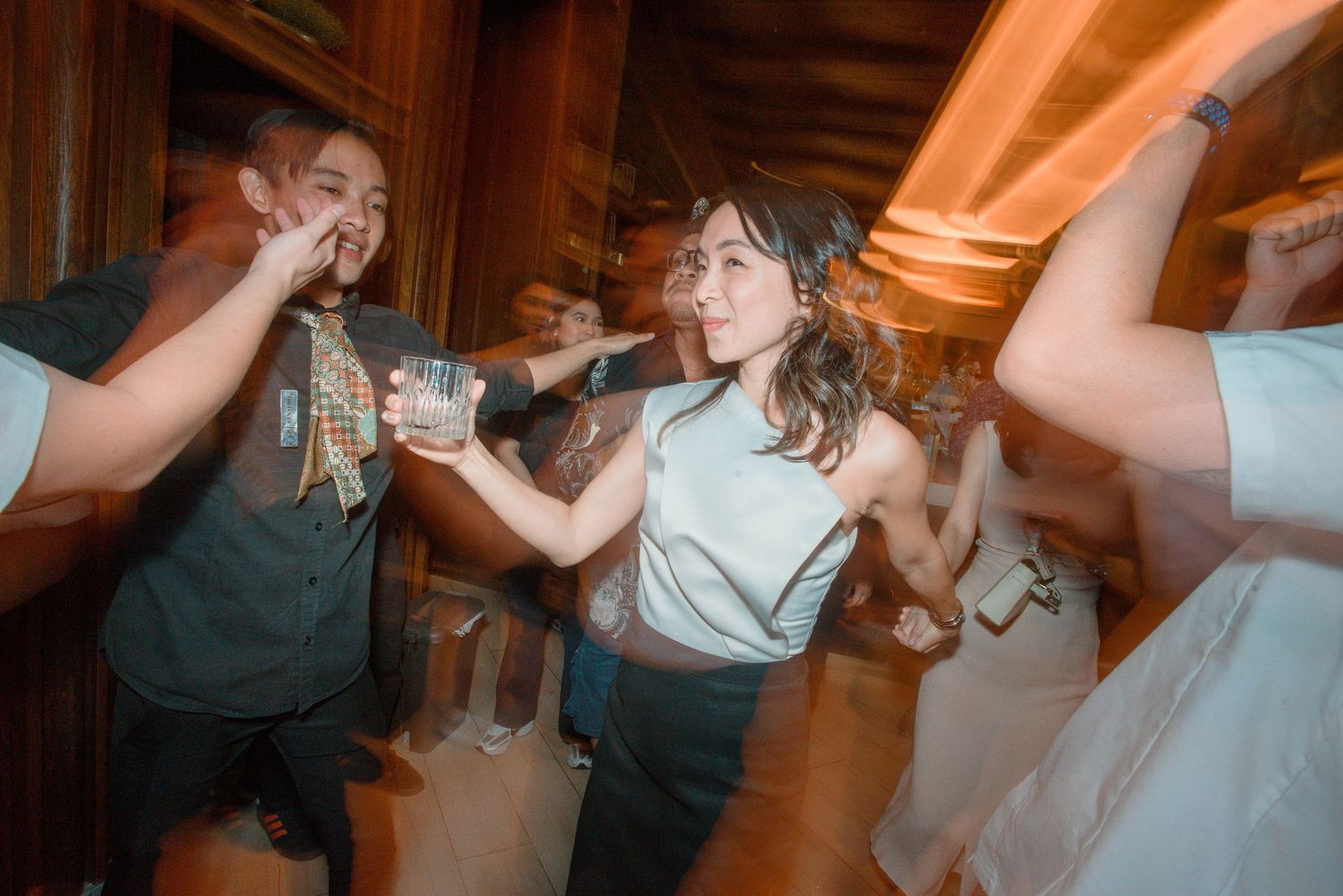 A group of people dancing and celebrating at a lively wedding reception with warm lighting and joyful atmosphere.
