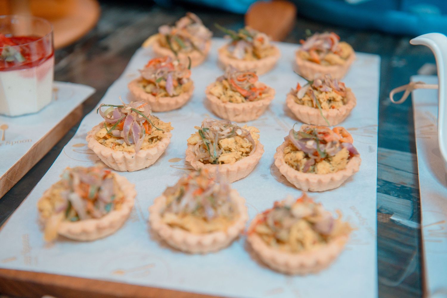 Rows of small pastry tarts filled with a creamy mixture and topped with nuts arranged on a white surface.