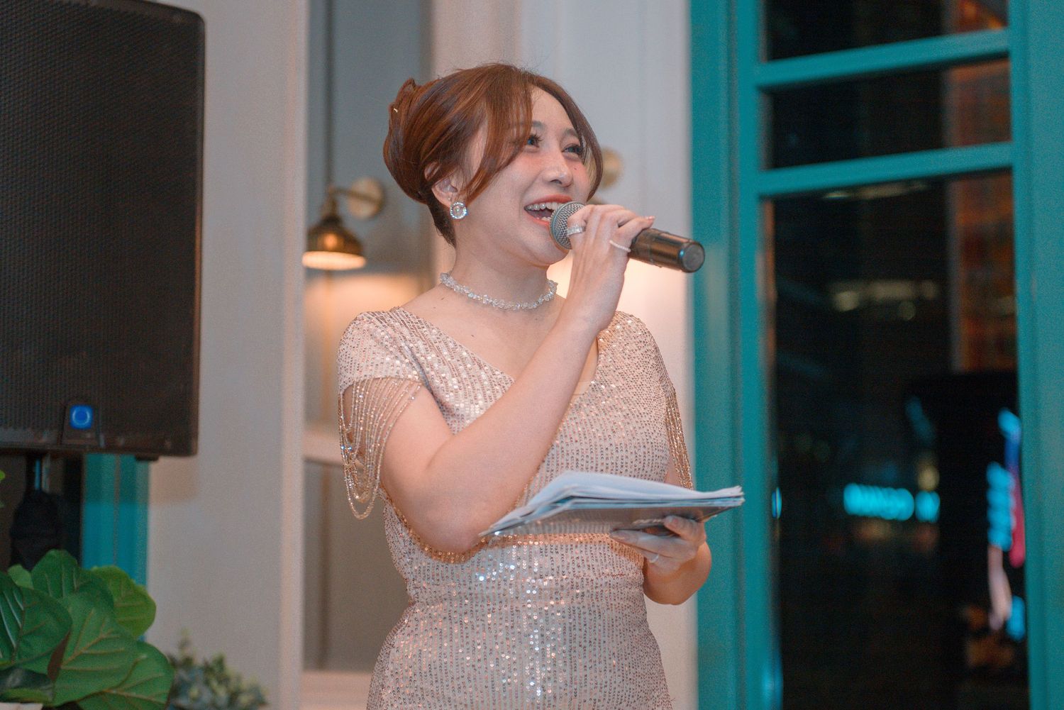 A person in a sparkly dress holds a microphone while singing at an event with blue lighting in the background.
