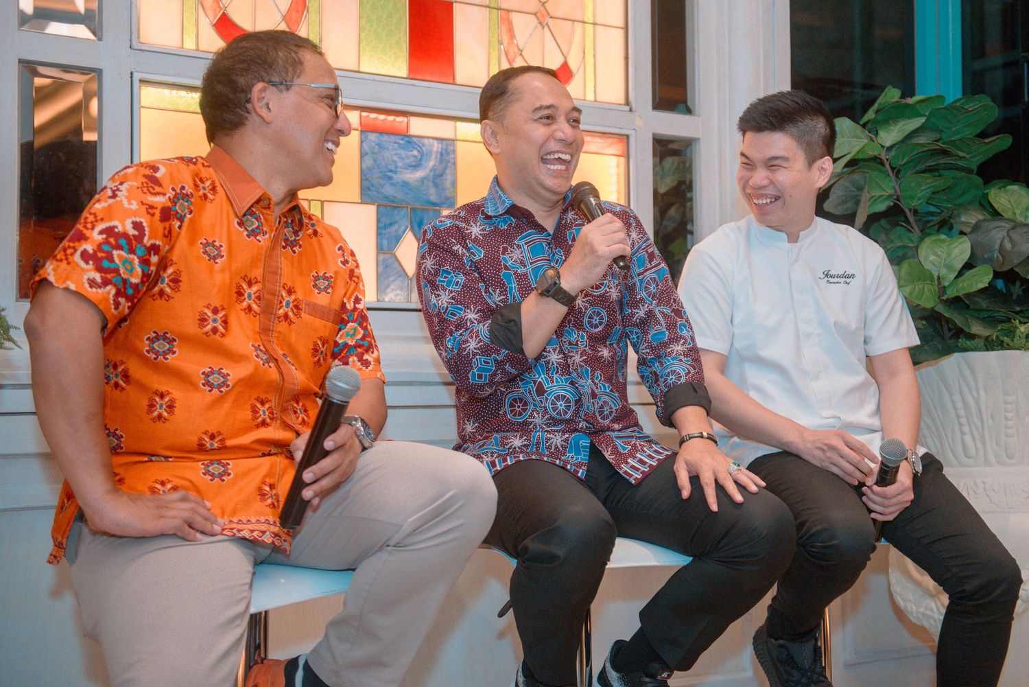 Three people sitting together and laughing while wearing colorful shirts in a casual indoor setting.