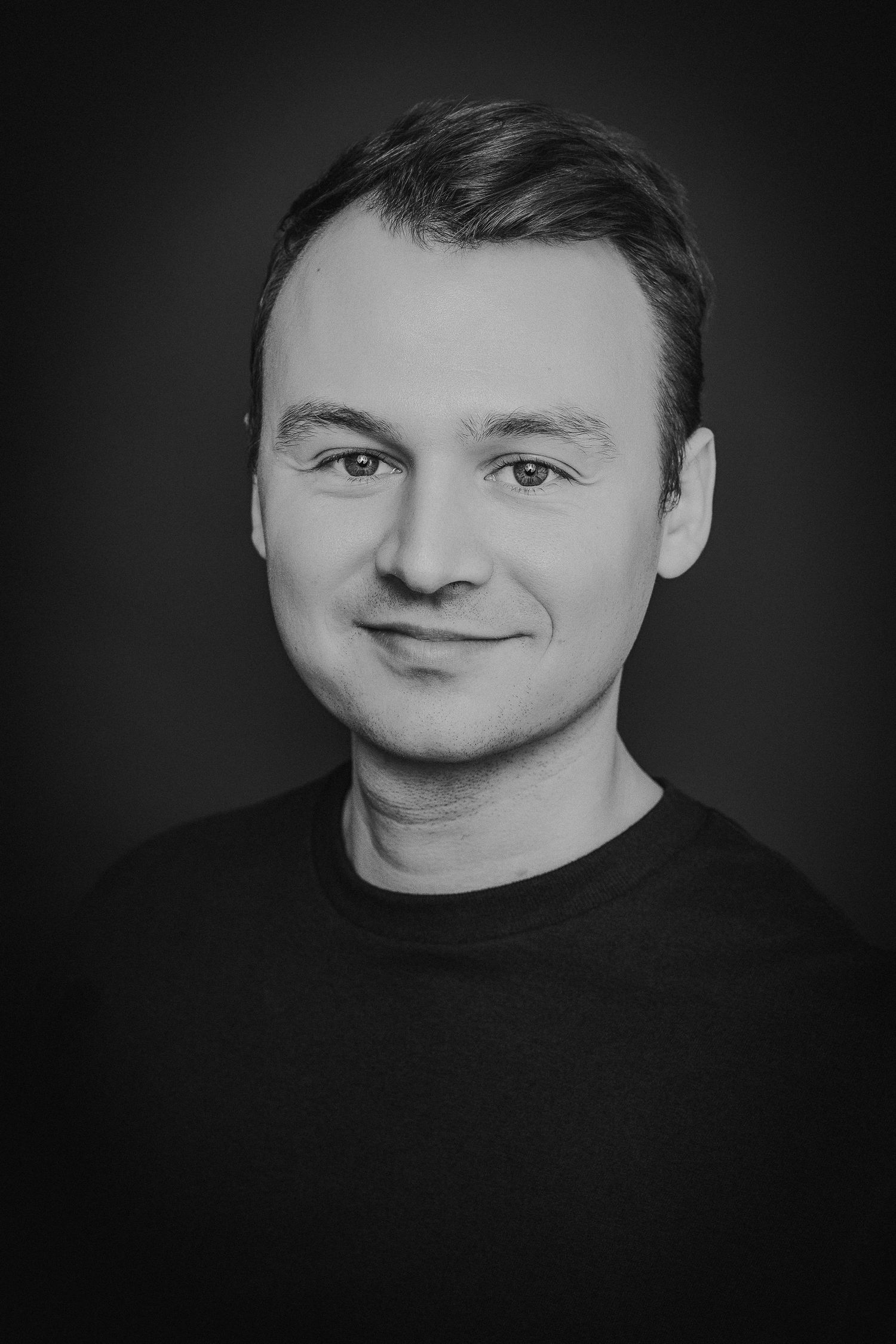 Professional black and white portrait photograph showing a person in a dark shirt against a dark background.