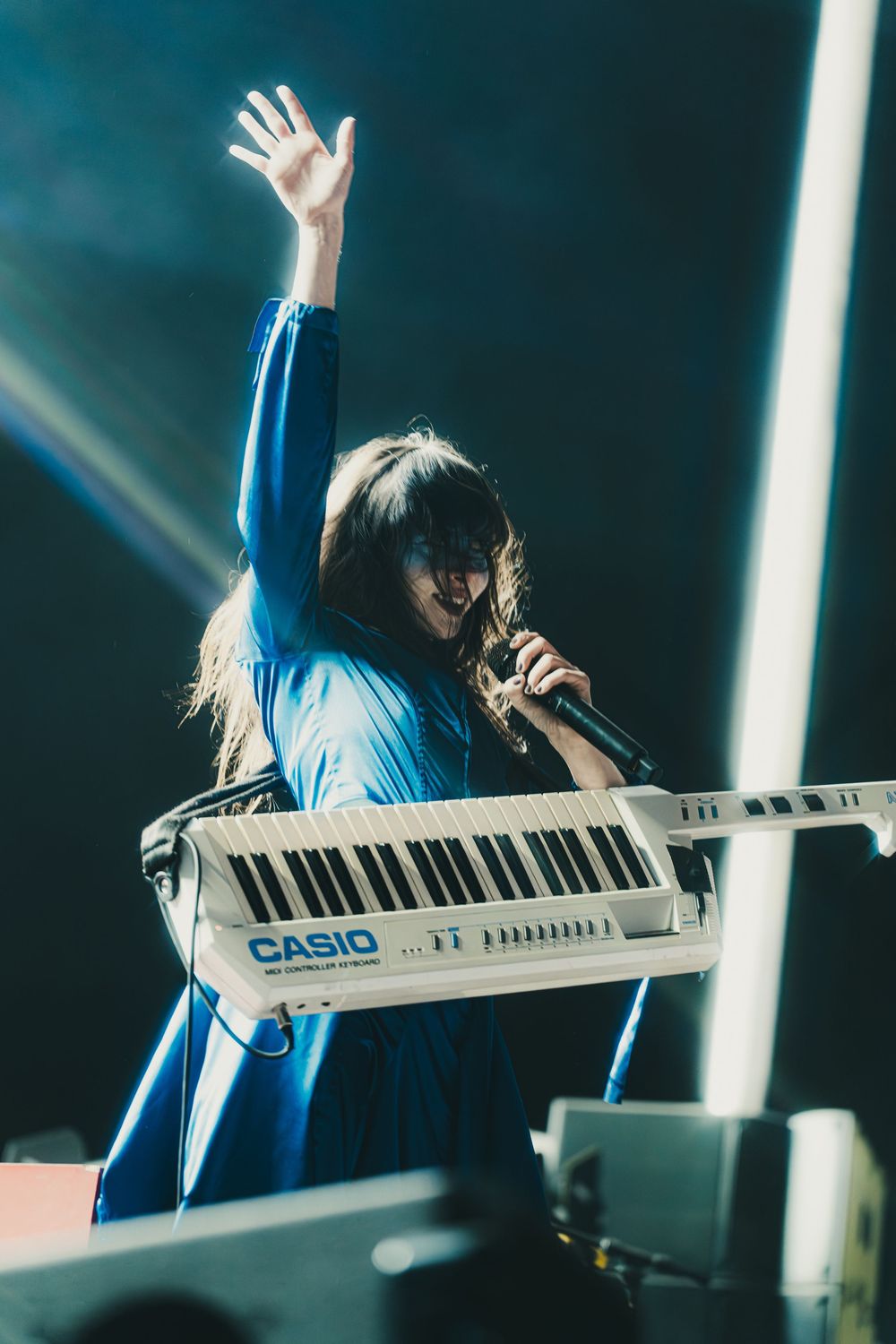 Musician in blue shirt performs energetically on a Casio keyboard during a concert with dramatic lighting.