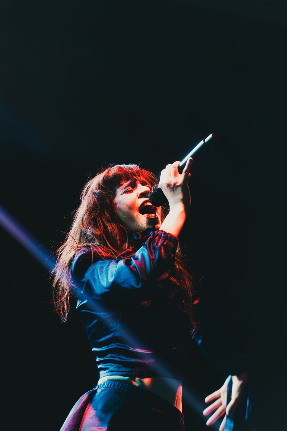 Energetic performer in blue outfit sings passionately on stage with dramatic lighting effects during live concert performance.