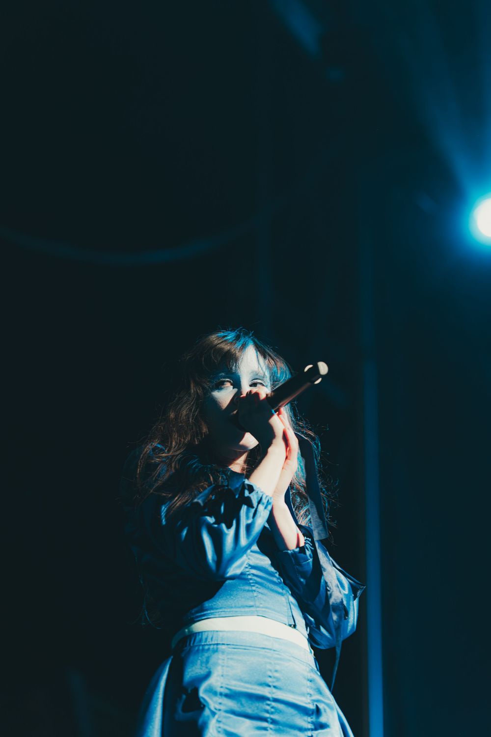 A performer in blue jeans sings passionately into a microphone on a dark stage with dramatic blue lighting.
