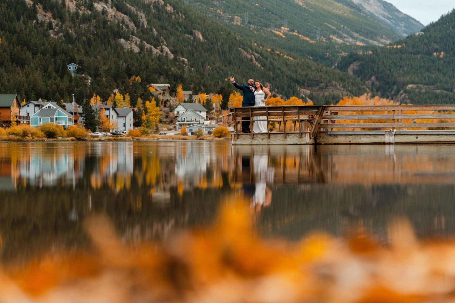 Couple on wooden dock overlooking reflective lake with autumn foliage and mountain town backdrop.