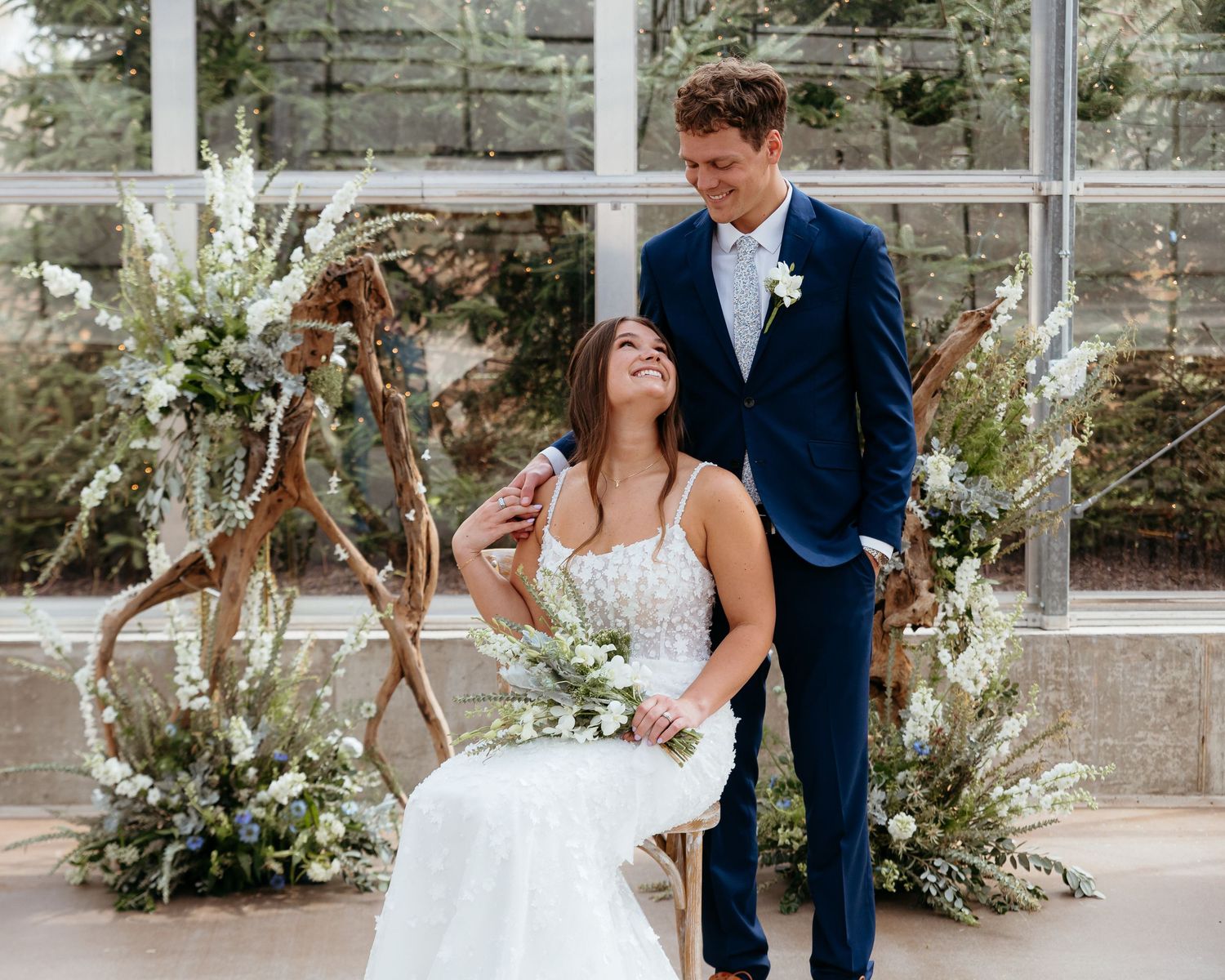 A bride and groom poses in front of a greenhouse with elegant floral wedding decorations. Ivy House ceremony space.