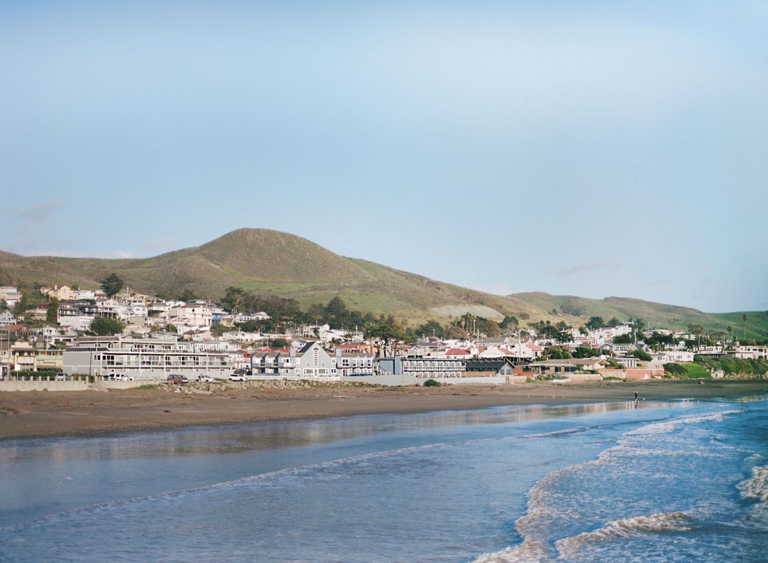 Panoramic view of Cayucos Beach and pier with coastal homes and rolling hills in the background.