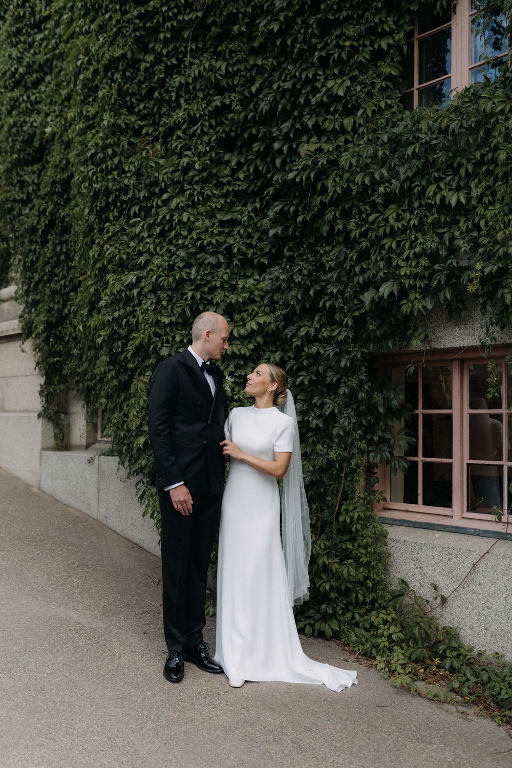 A couple in wedding attire stands together against an ivy-covered wall of a building.