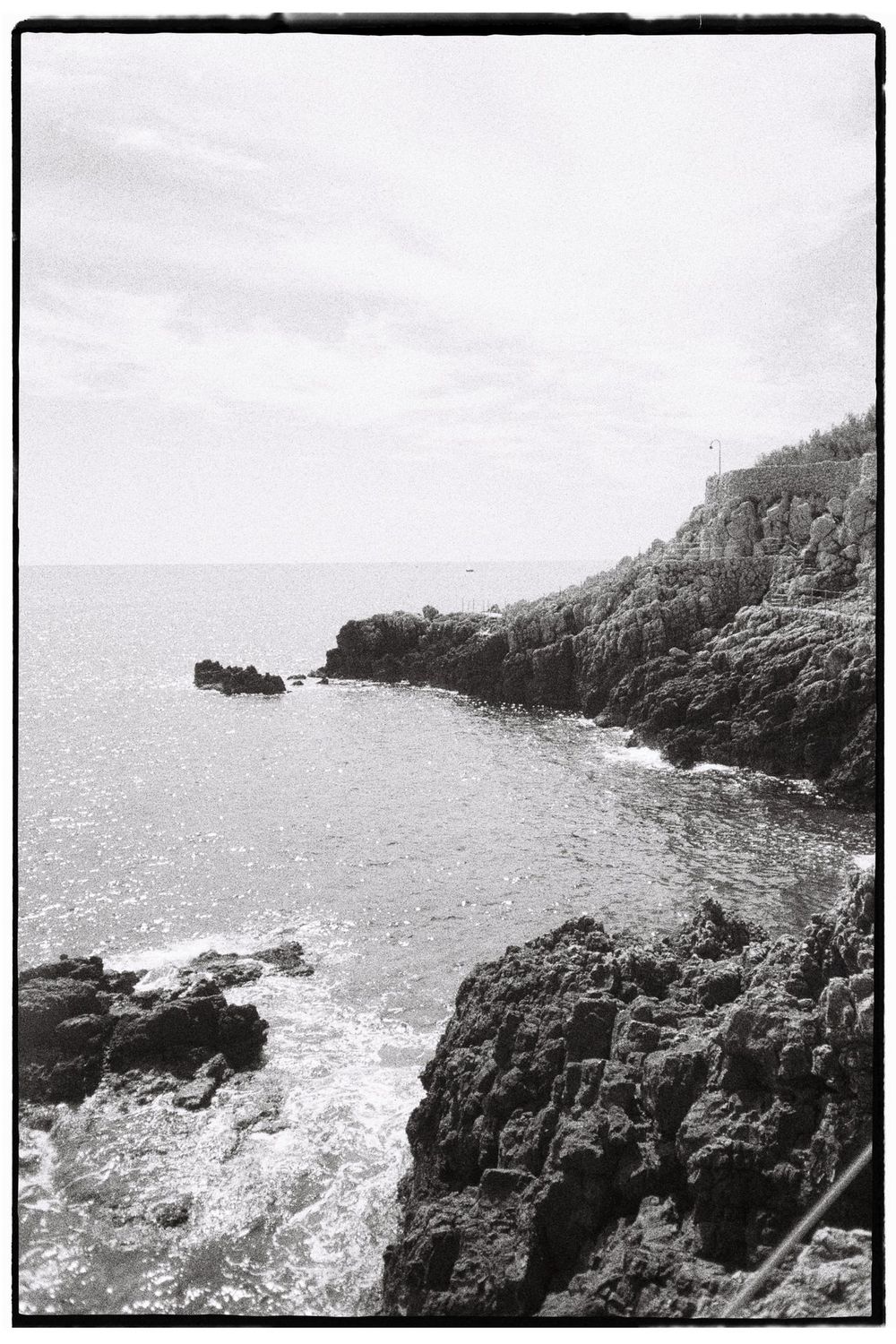 A black and white image showing a rocky coastline with a body of water in the foreground.