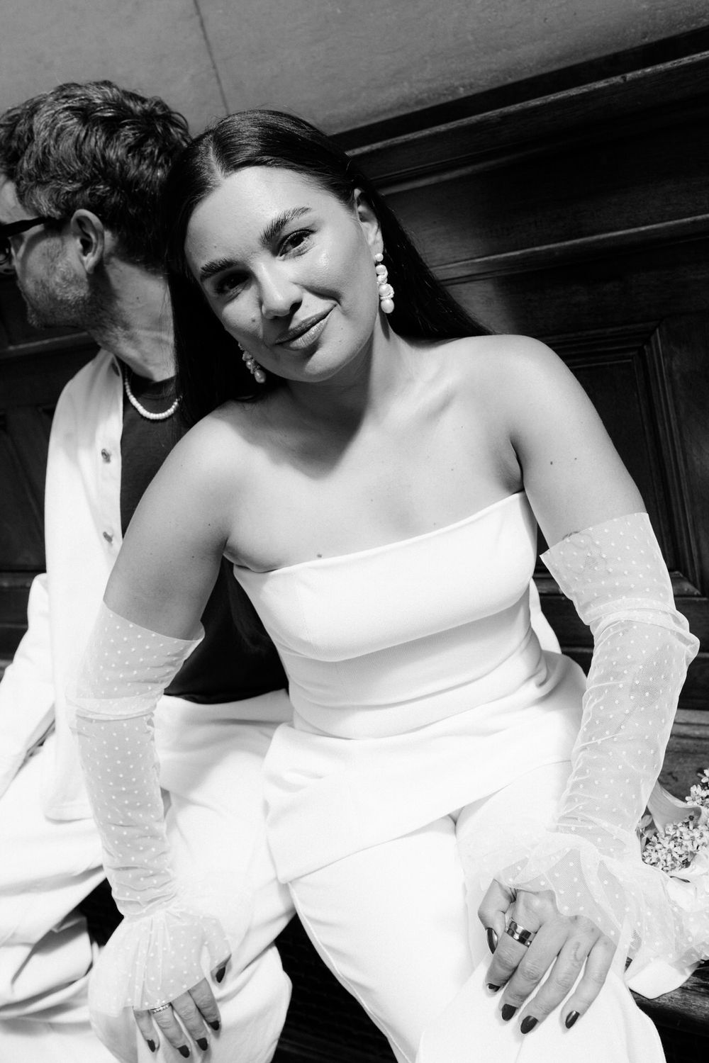 Black and white photo of a smiling woman in a white strapless outfit with lace gloves at a formal event.