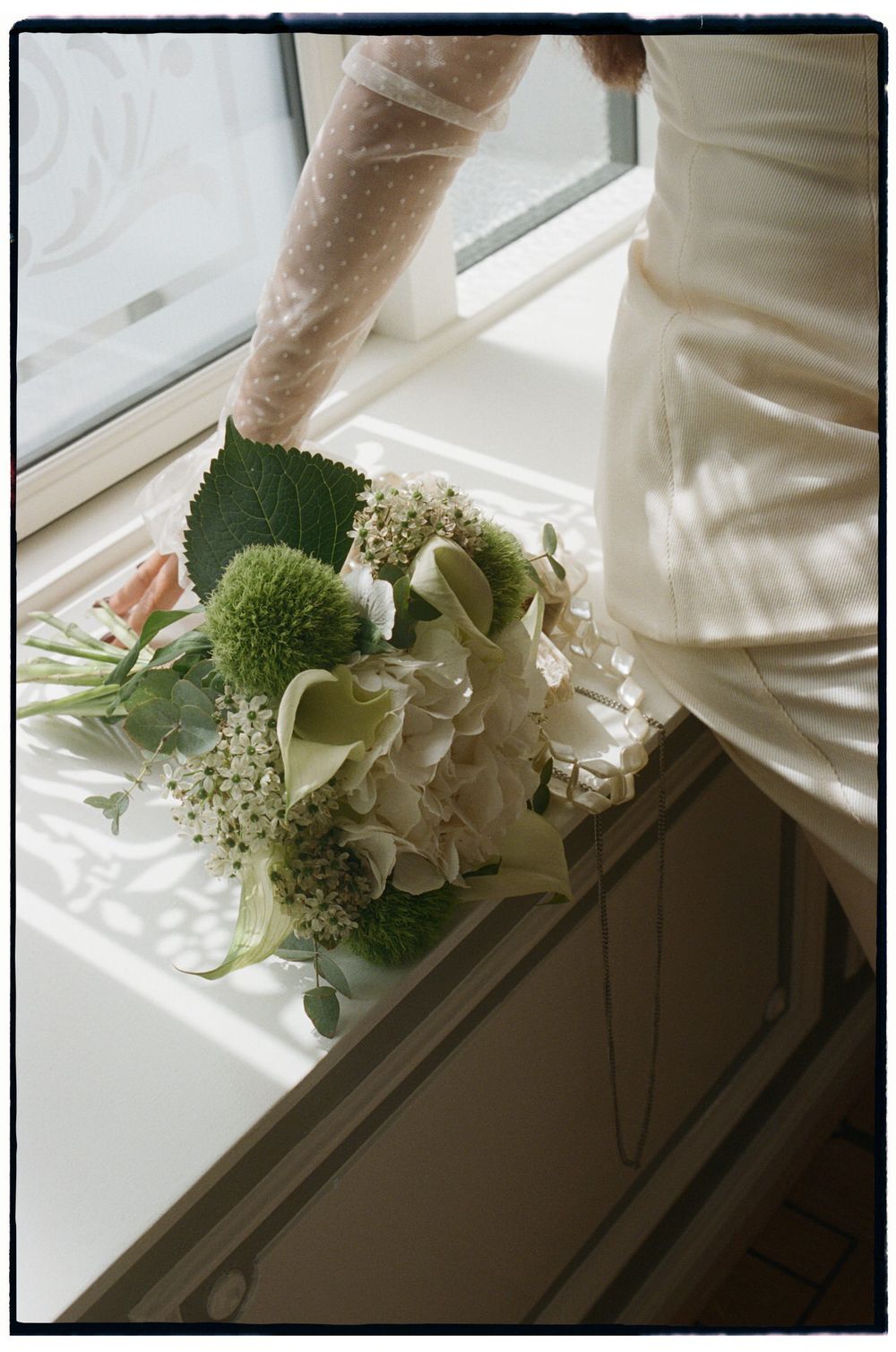 Bride holding a white and green wedding bouquet with hydrangeas near a sunlit window in a white dress.