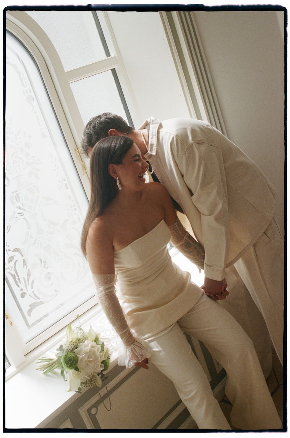 Bride and groom share a romantic kiss by a sunlit window, she in a strapless white gown holding a bouquet.