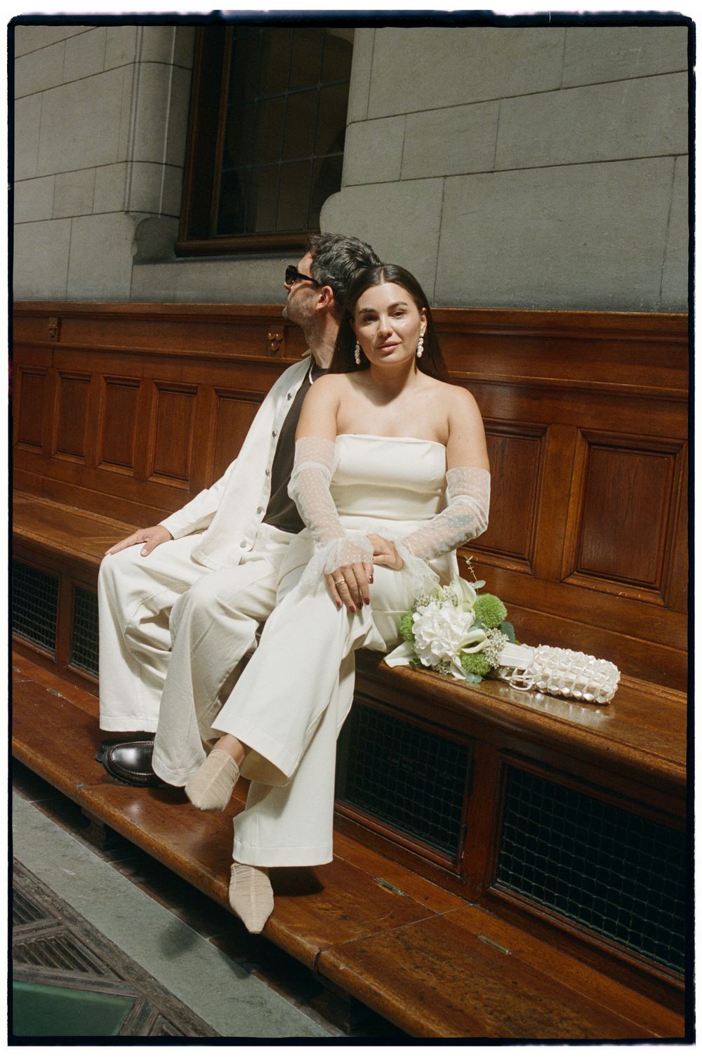Couple in white wedding attire sitting on wooden bench, bride in strapless jumpsuit with sheer gloves holding floral bouquet.