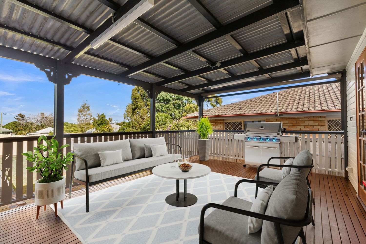 Spacious outdoor entertaining deck with grey sofa set, BBQ, pergola roof, and leafy garden views.