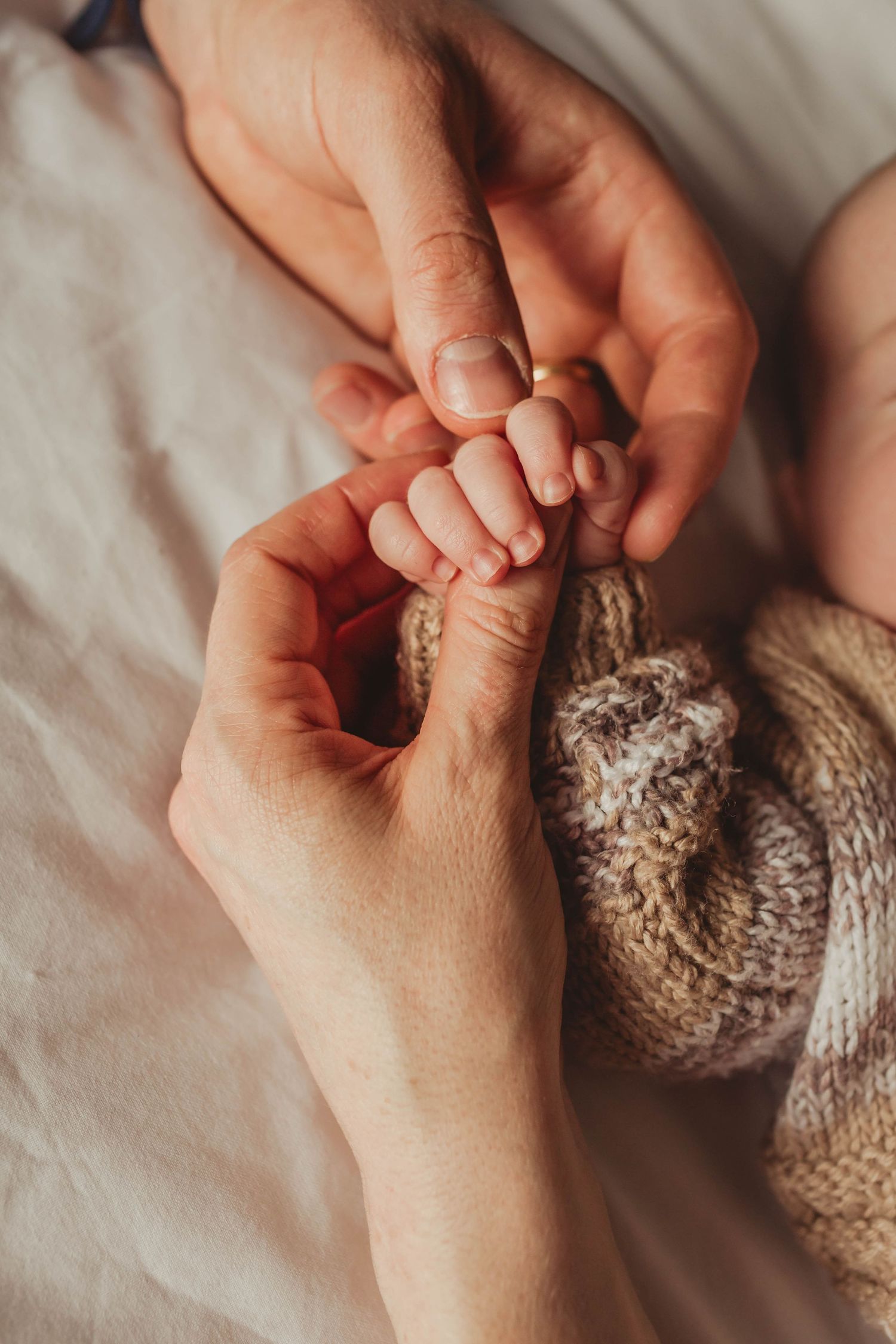 Close up of gentle hands holding tiny baby feet with knitted blanket on white bedding.