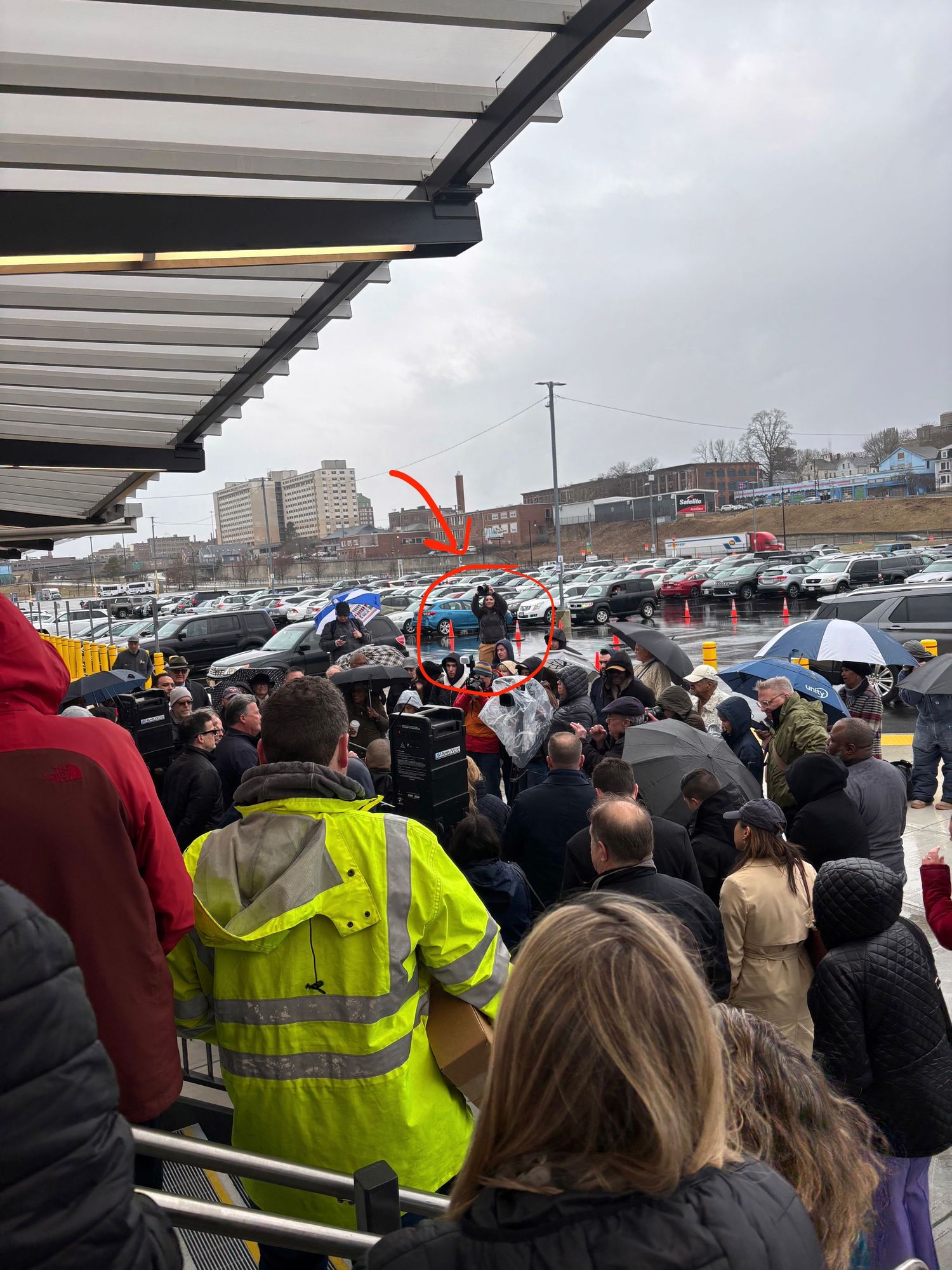 A large crowd gathers under covered walkways in a parking lot during what appears to be a rainy day event.