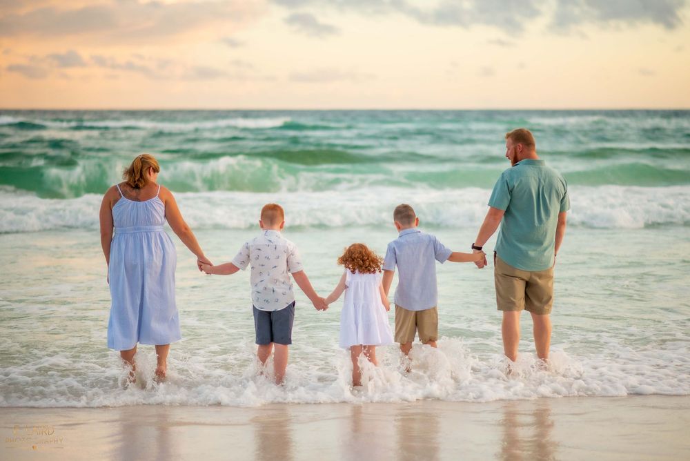 Family holding hands while walking along beach shoreline at sunset with waves crashing behind them.