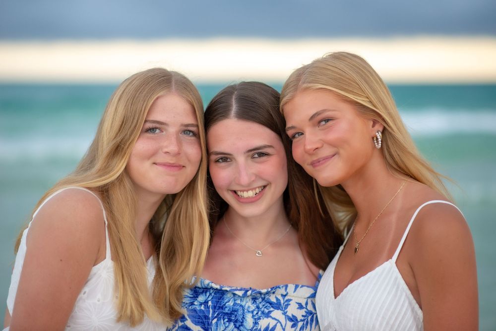 Three friends in blue and white dresses pose together on a sunny beach with ocean waves in the background.