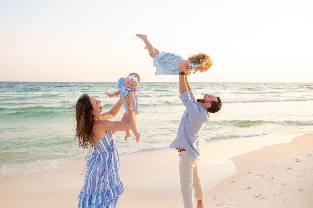 A couple in blue and white outfits dances playfully together on a sunny beach with waves in the background.